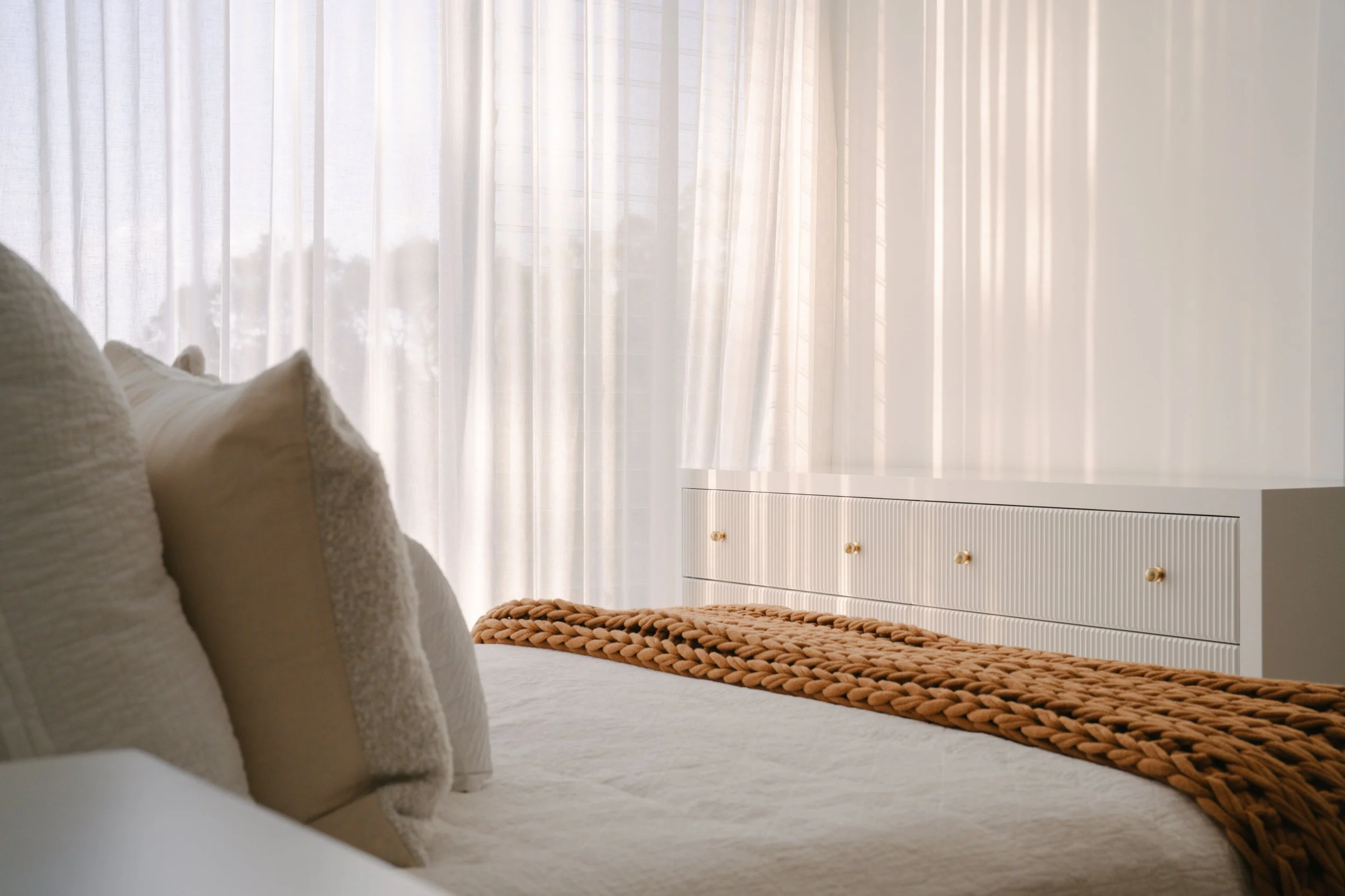 A cozy bed with beige and white pillows, a brown woven blanket, and white curtains letting in natural light, with a white dresser in the background.