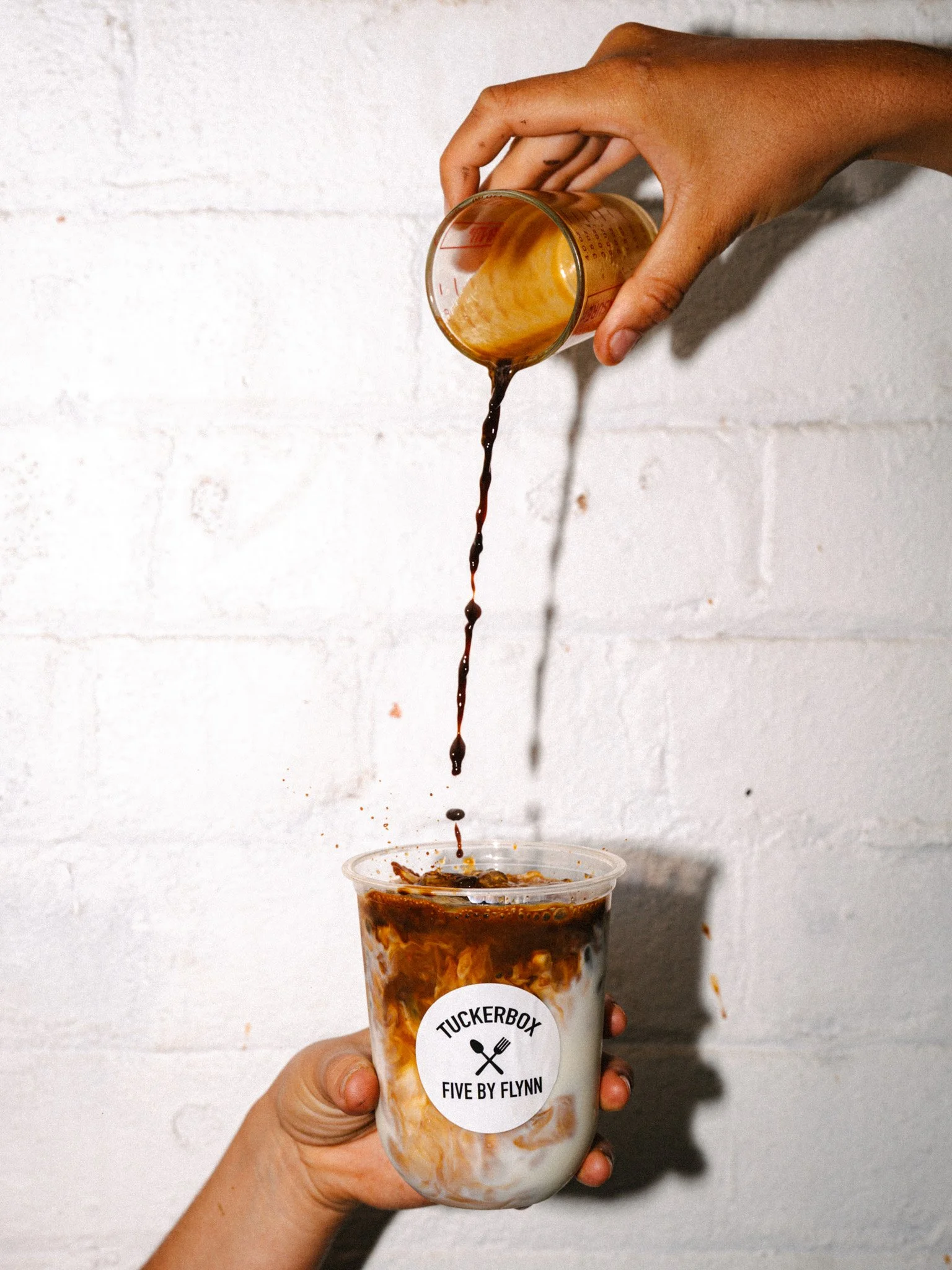 Person pouring coffee syrup into a cup with milk, which has a label reading "Tuckerbox, Five by Flynn".