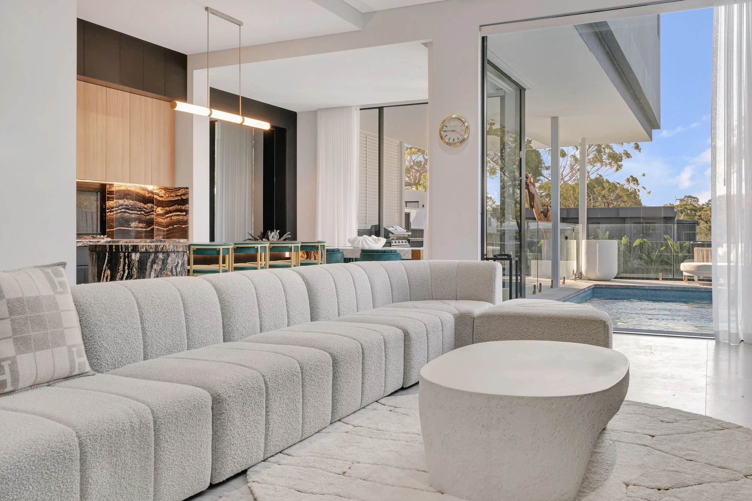 Modern living room with a light gray sectional sofa, textured throw pillow, round coffee table, and a view of a pool outside through glass sliding doors.