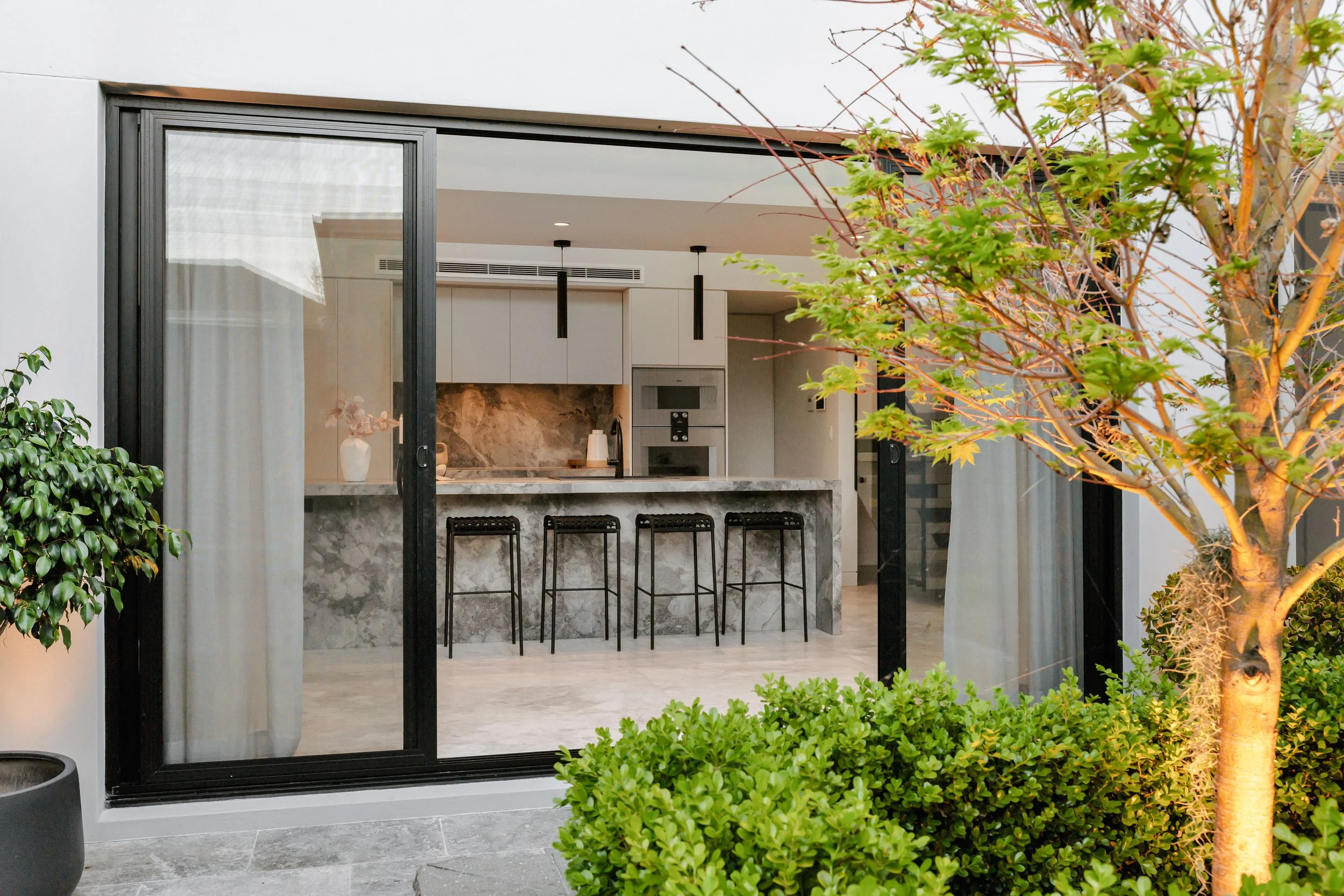 View of a modern indoor kitchen seen through sliding glass doors, with a marble kitchen island, black bar stools, white cabinets, and a marble backsplash, surrounded by green plants and trees outdoors.