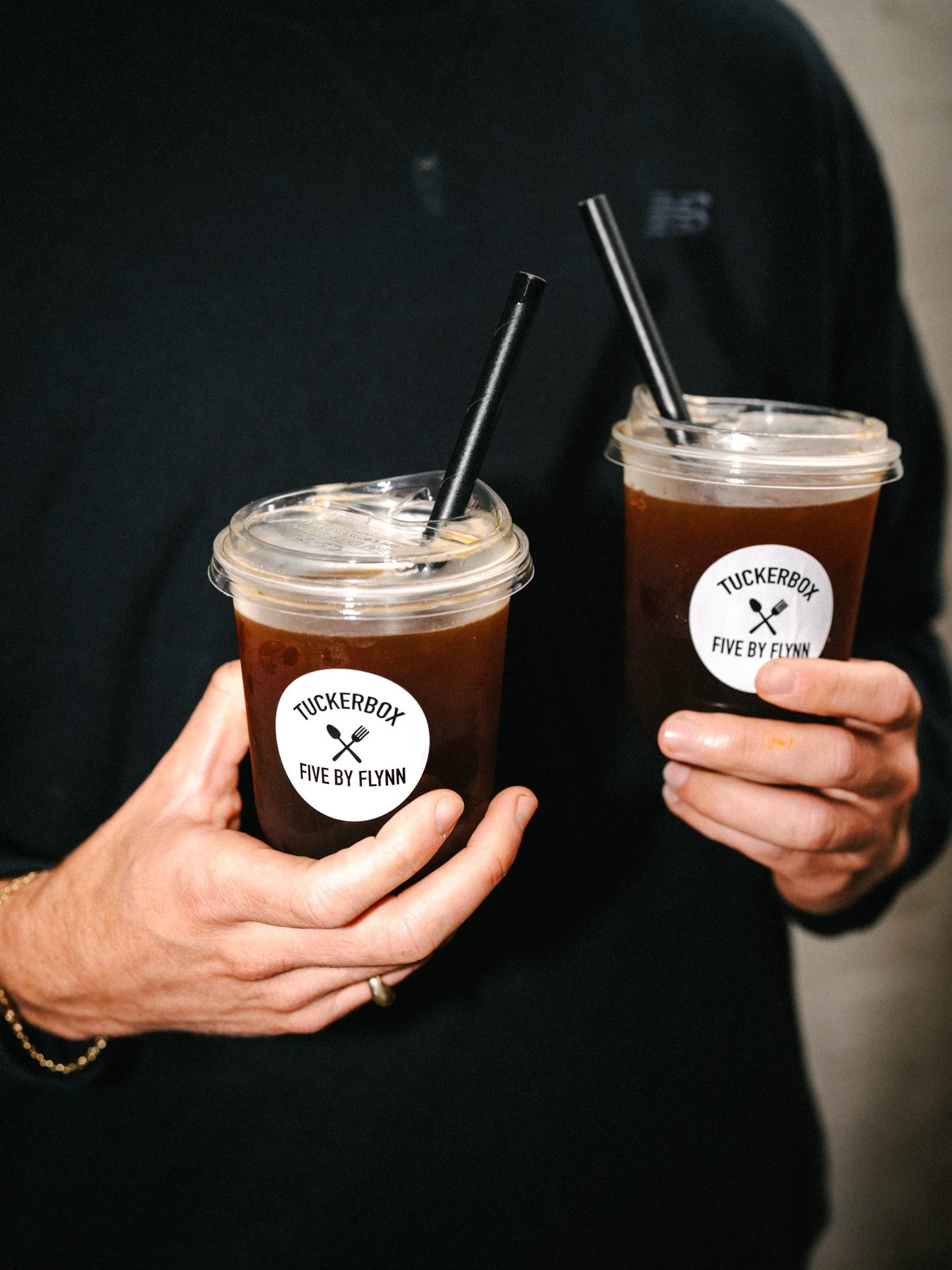 Two hands holding plastic cups with dark iced drinks, labeled 'TUCKERBOX FIVE BY FLYNN', with black straws, against a dark background.