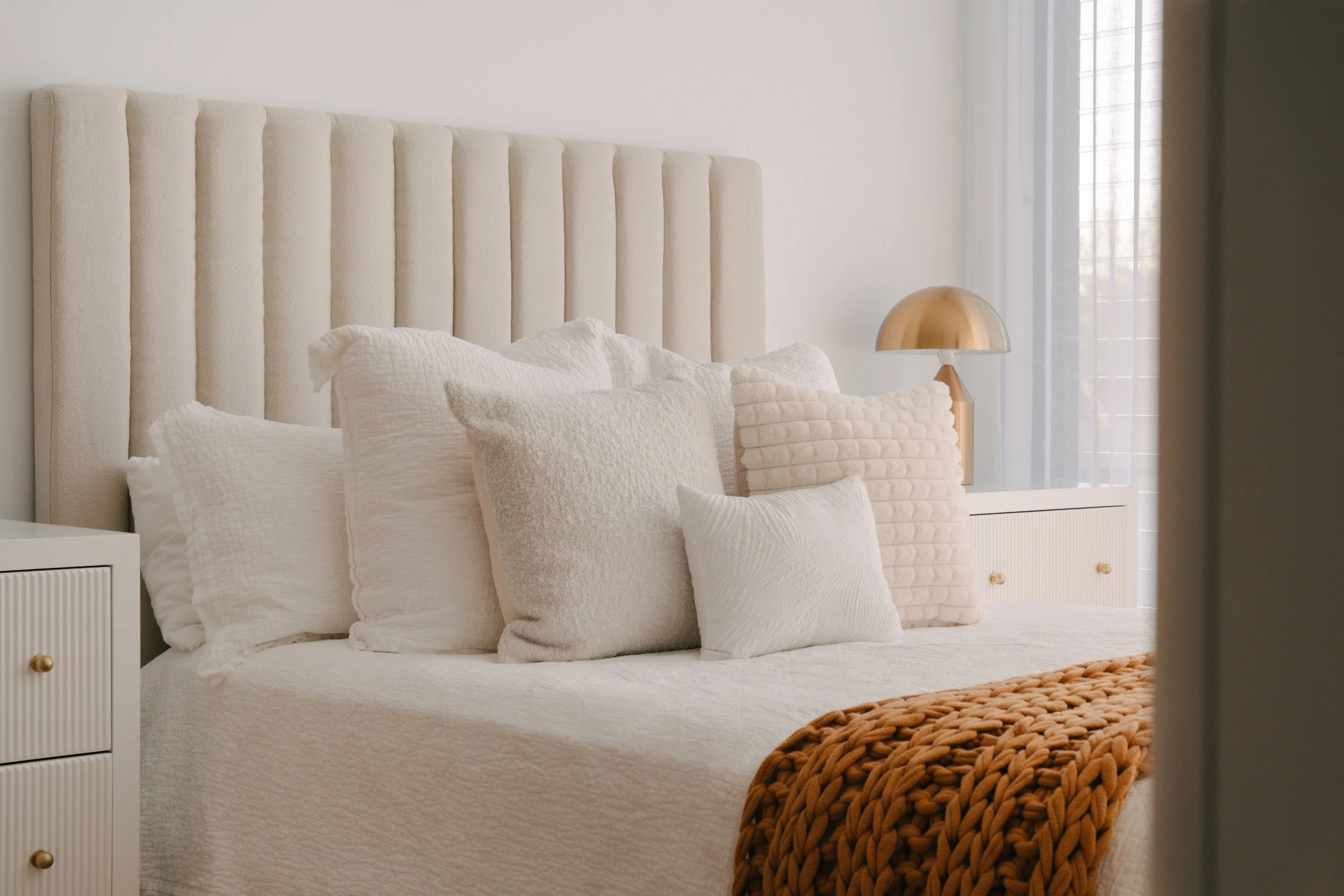 A neatly made bed with a cream-colored headboard, multiple white and beige pillows, and a chunky knit brown blanket at the foot. There are white nightstands with gold knobs on each side, and a gold table lamp on the right nightstand. Curtain-covered
