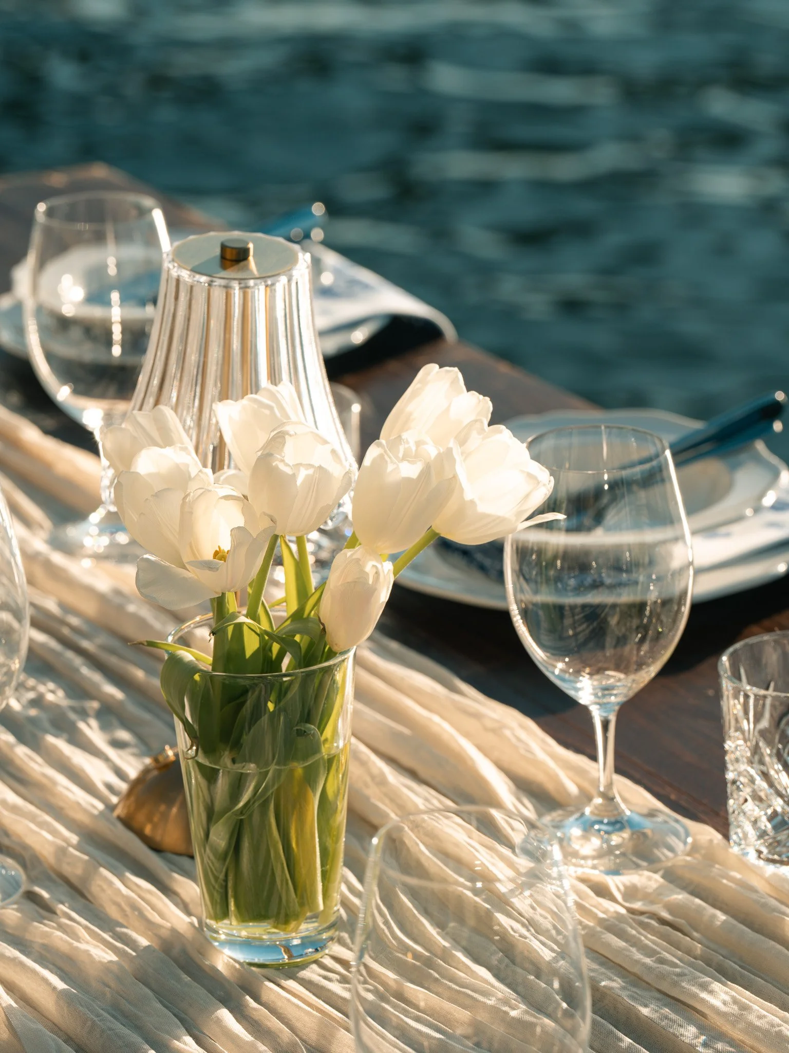 A table set with white tulips in a glass vase, surrounded by glassware and a lamp, with water in the background.