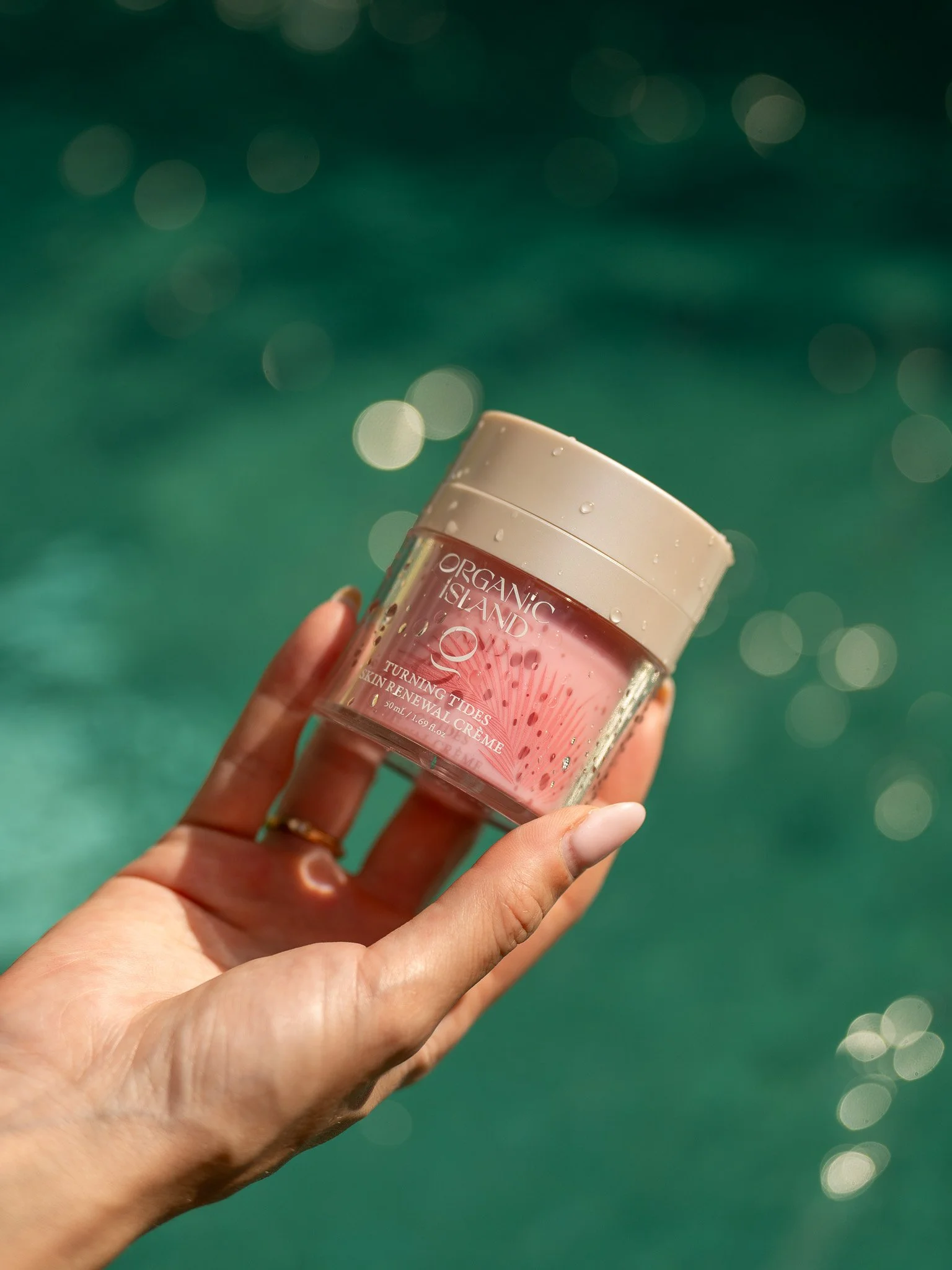 Hand holding a jar of Organic Island skin renewal creme against a blurred ocean background with sunlight reflection.