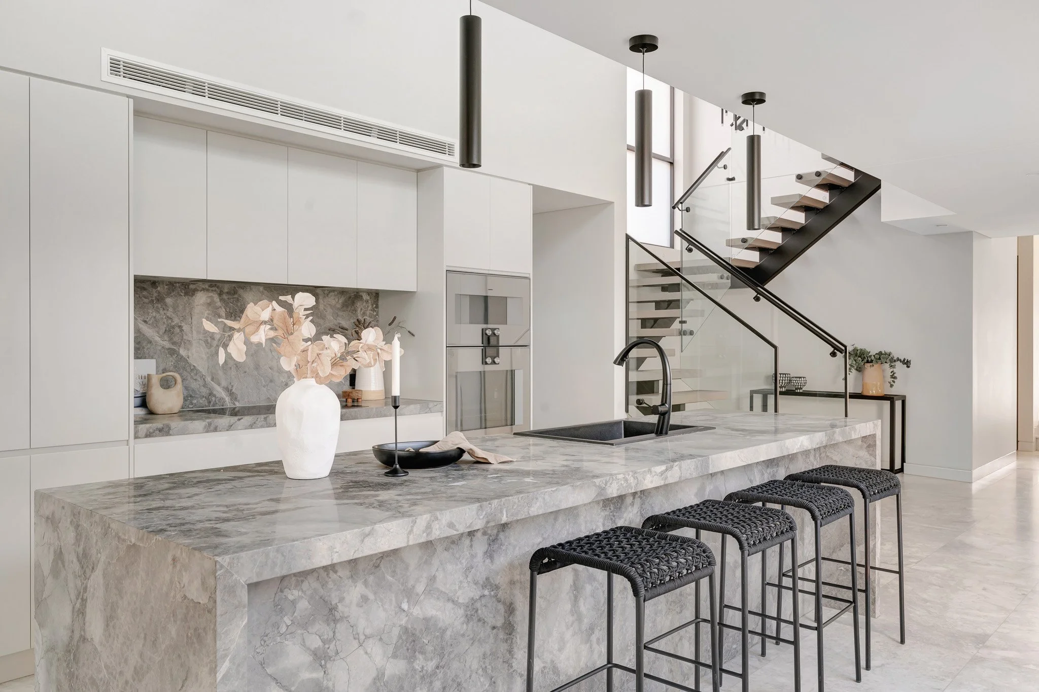 Modern kitchen with marble island, white cabinetry, black fixtures, and a staircase with glass railing in the background.