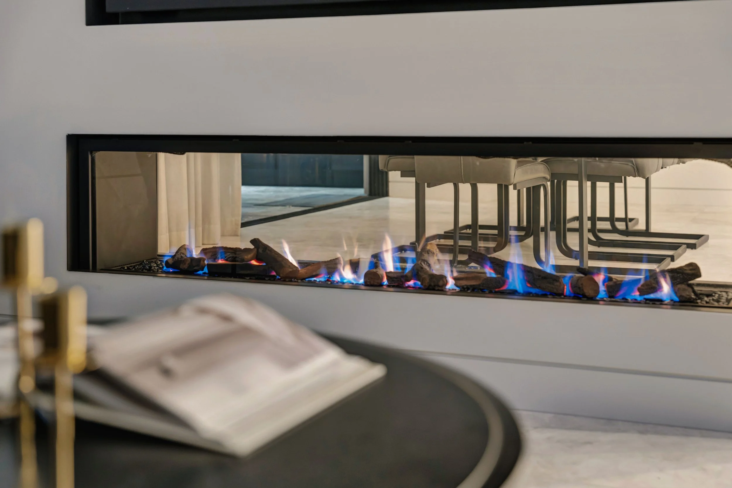 Modern fireplace with blue flames and logs, seen through a rectangular glass window in a white wall, with a dining table with beige chairs reflected in the background.