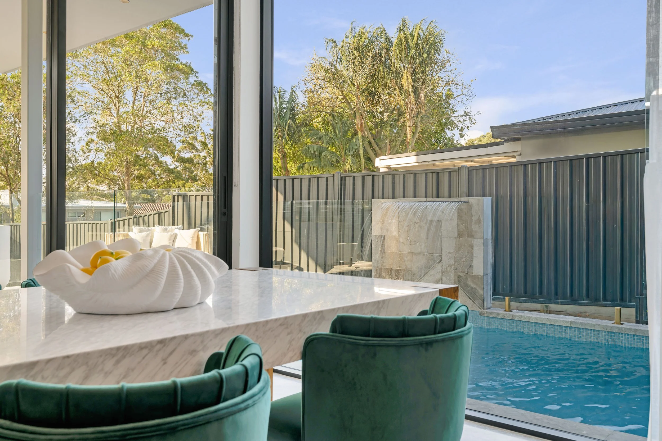 Indoor dining area with green velvet chairs and a marble-topped table, overlooking a backyard with a small pool, a water feature, and lush green trees outside large glass sliding doors.