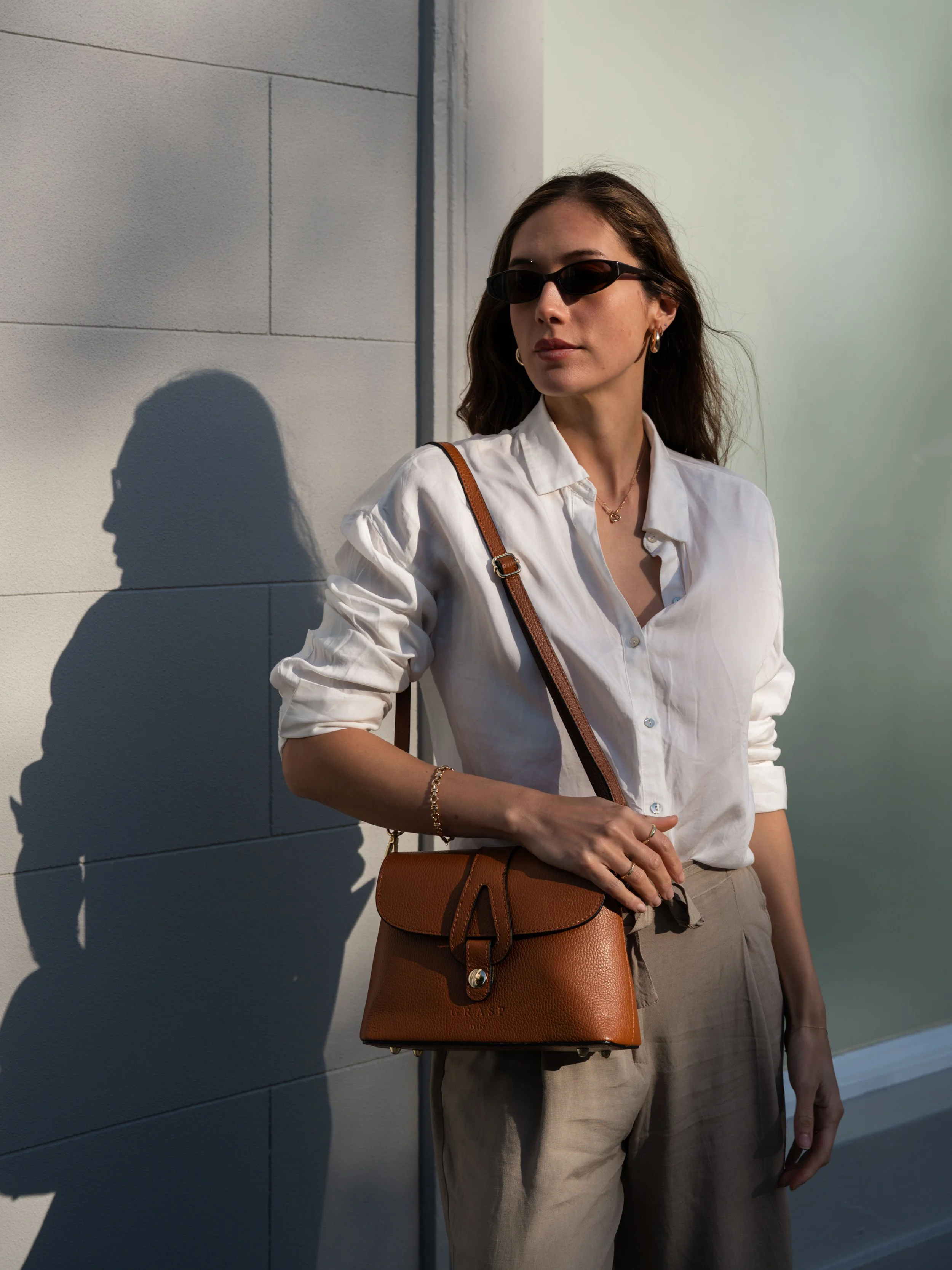 A woman standing outdoors against a light-colored wall, wearing black sunglasses, a white button-up shirt with rolled-up sleeves, beige pants, and carrying a brown leather handbag with a long strap, illuminated by sunlight casting a shadow on the wal