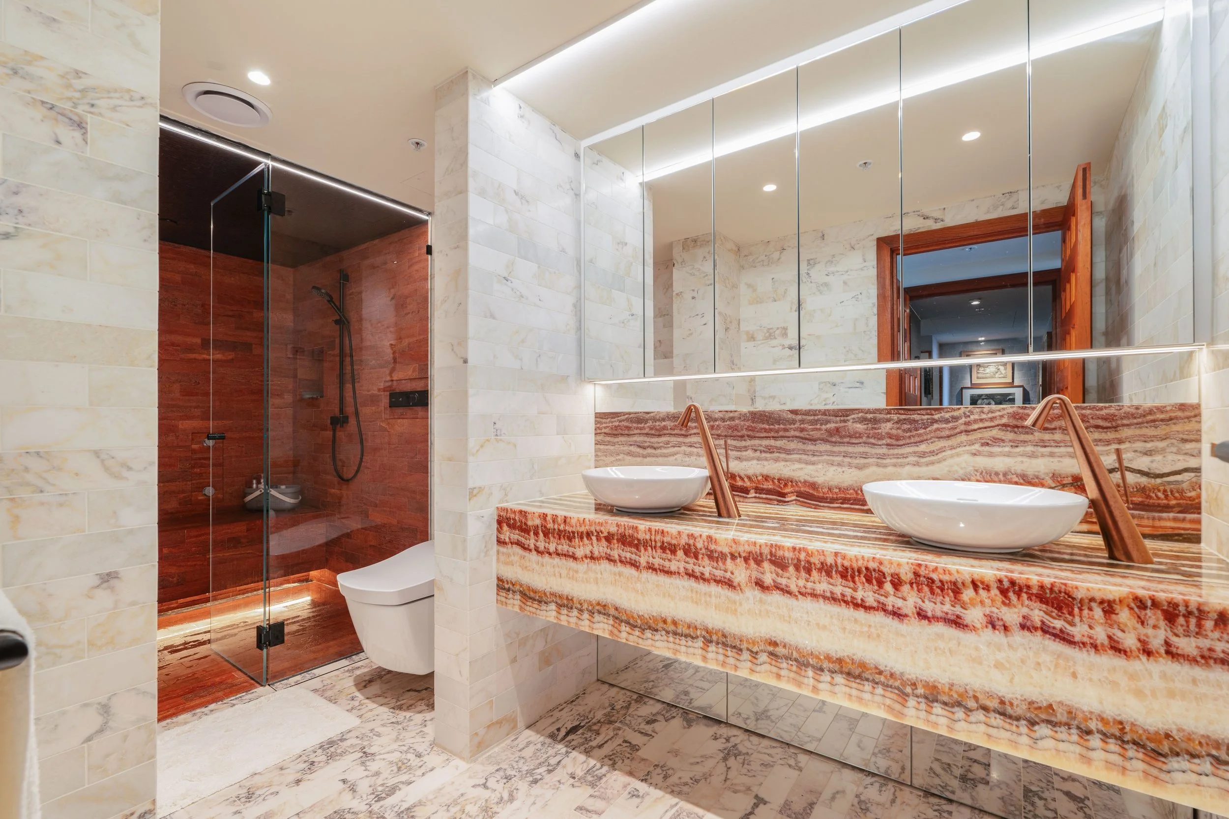 Modern bathroom with a double sink vanity, large mirror, marble and onyx surfaces, walk-in shower with glass door, and a wooden door leading to another room.