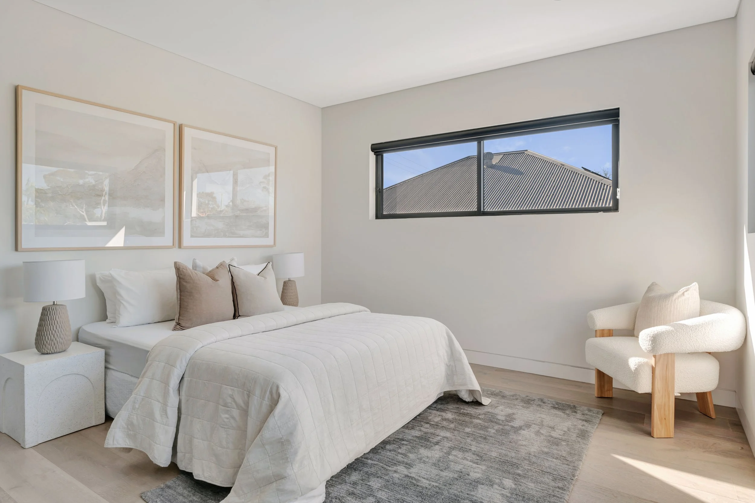Bright, minimalistic bedroom with a white bed, beige pillows, white nightstands with lamps, a textured rug, a cream armchair with wooden legs, and a large horizontal window showing a blue sky and a rooftop.