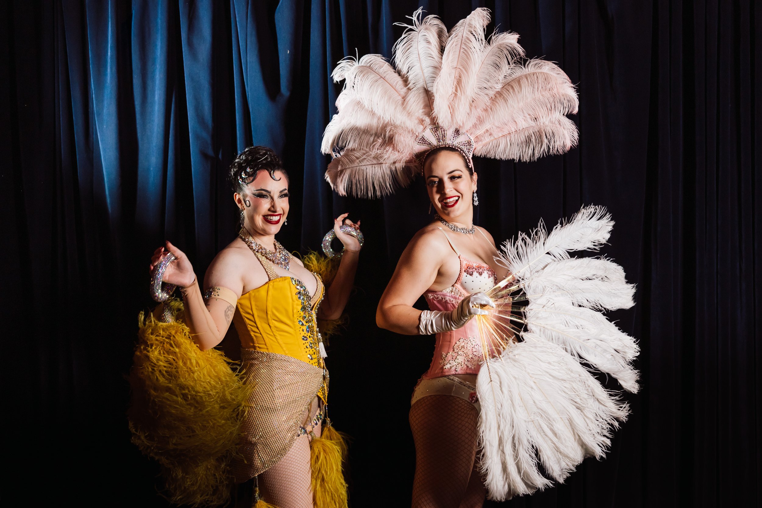 Two women dressed in vibrant burlesque costumes with feathers, sequins, and jewelry, standing on a dark stage with black curtains, smiling and posing.