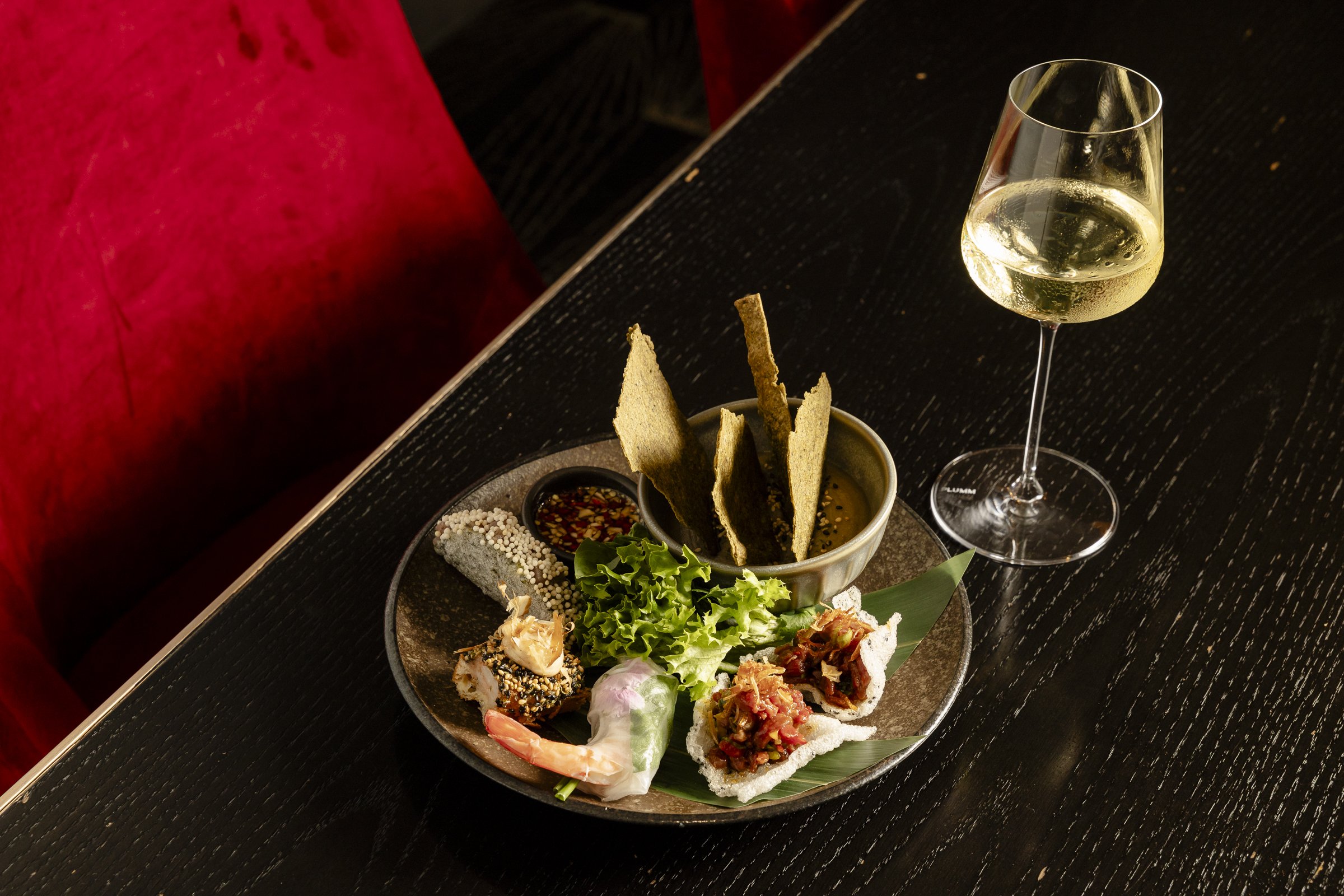 A black plate with various sushi pieces, garnishes, and sauces, accompanied by a glass of white wine on a black textured table.