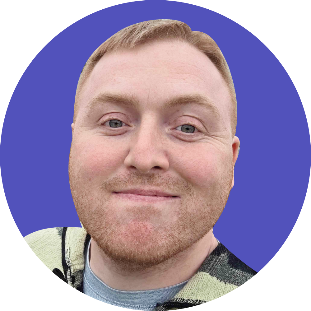 Close-up of a smiling man with red hair and a beard, wearing a light-colored hoodie, against a solid purple background.