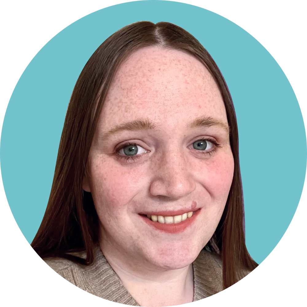 A woman with long red hair, blue eyes, and fair skin, smiling and wearing a beige sweater.