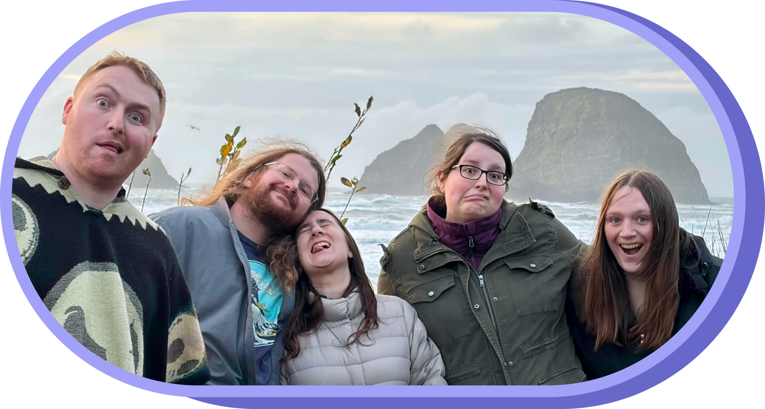 Group of five people making funny faces on a beach with rocks and ocean in background.