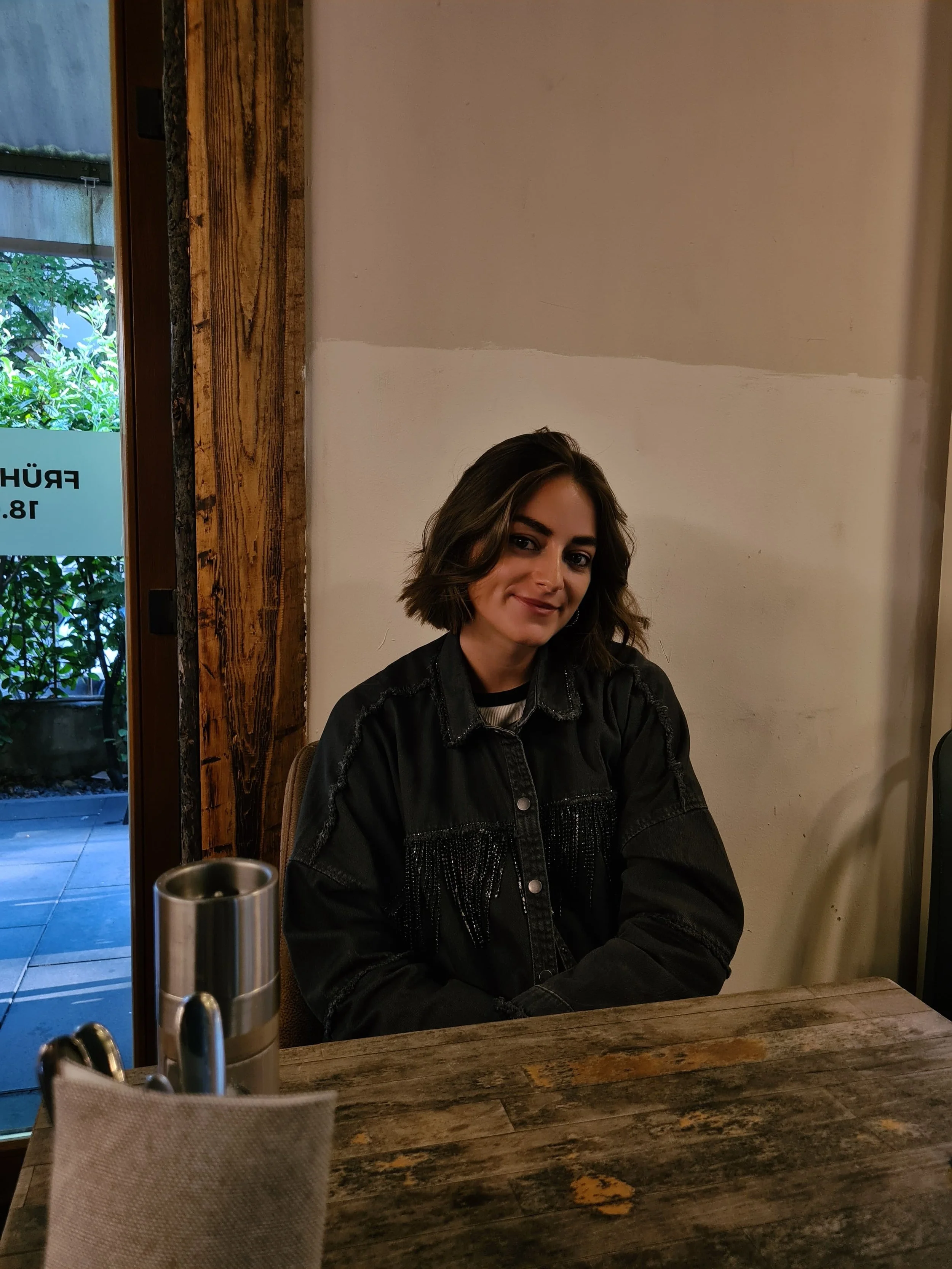 Junge Frau mit braunen, schulterlangen Haaren sitzt an einem Holztisch in einem Café, trägt eine schwarze Jacke und lächelt leicht.