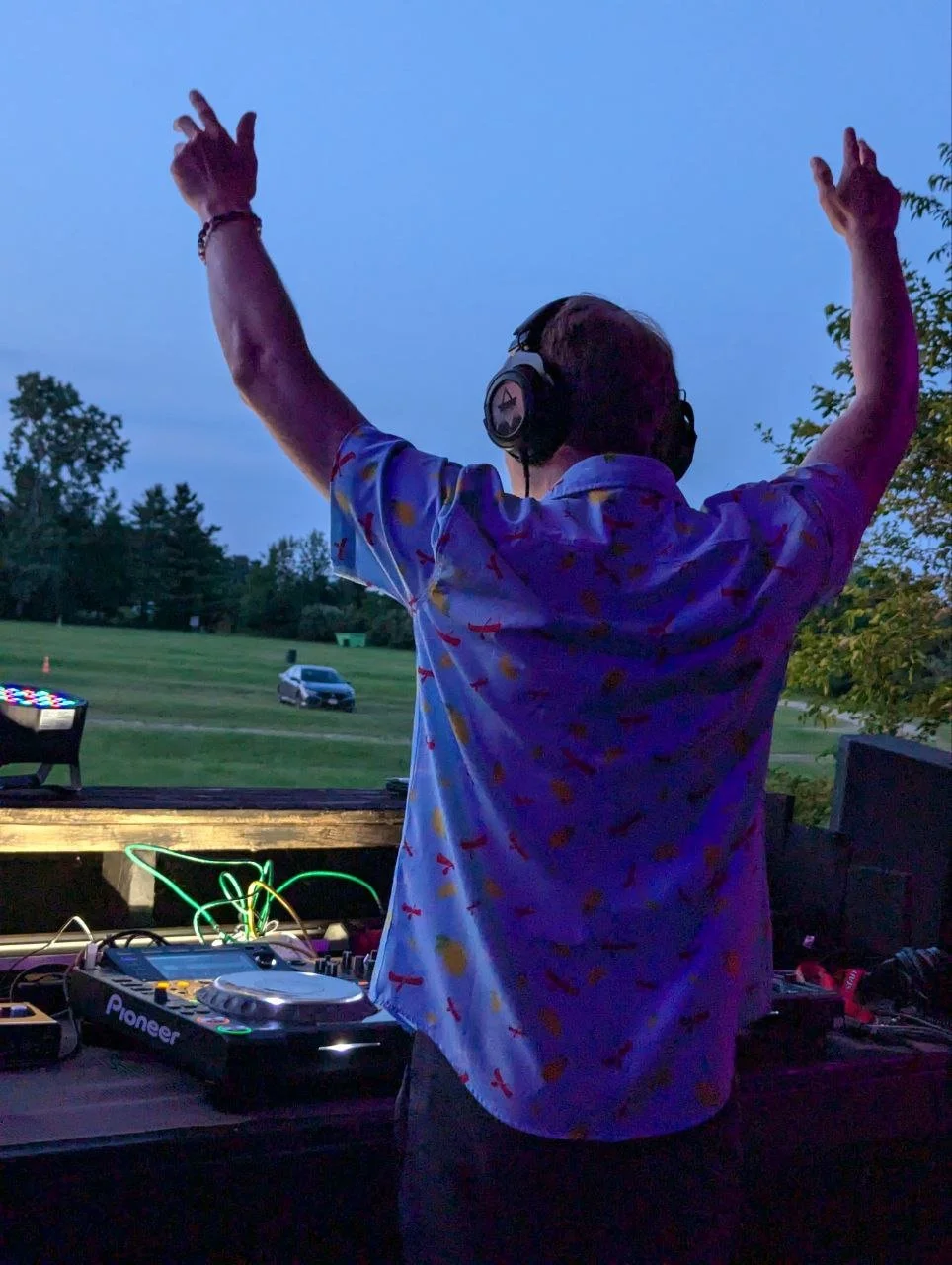 DJ with headphones wearing a colorful shirt, raising hands while standing at an outdoor DJ setup during dusk, with a field and trees in the background.