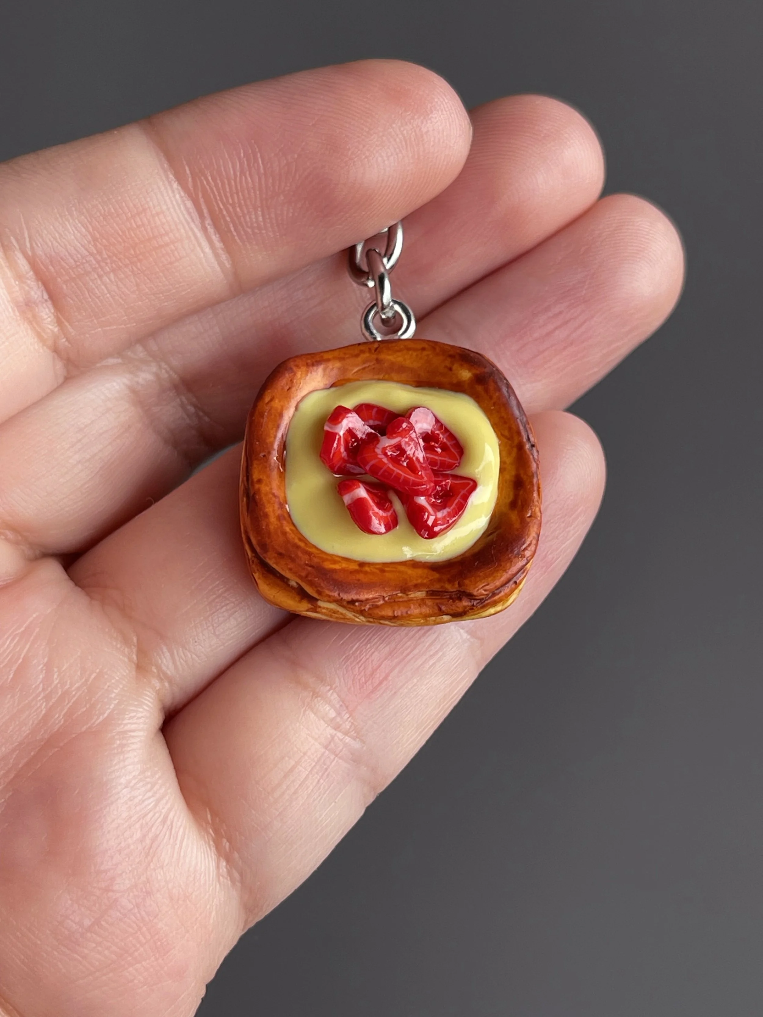 A hand holding a miniature danish pastry with yellow custard and strawberry slices keychain.