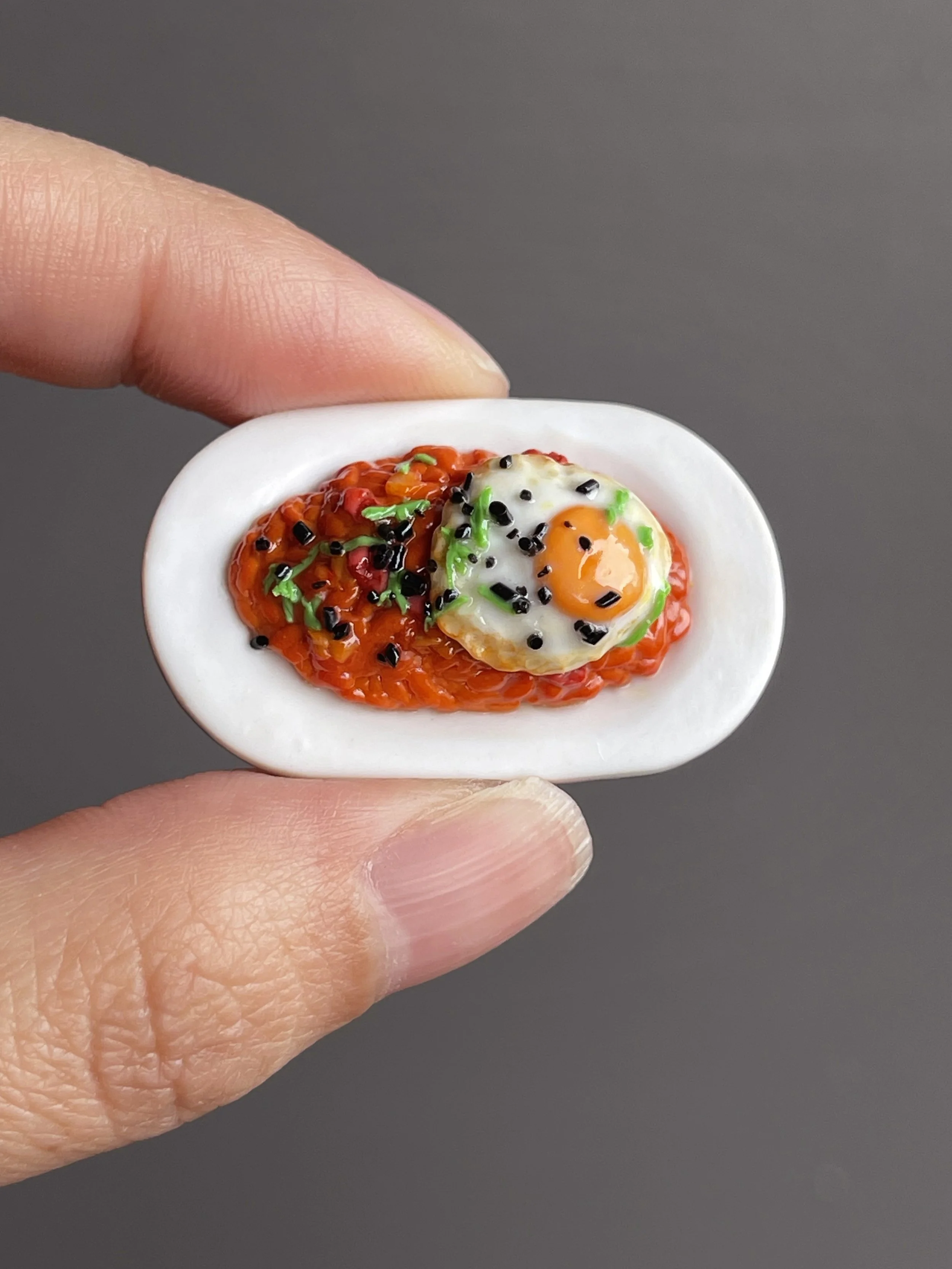 Fingers holding a miniature polymer clay kimchi fried rice topped with a fried egg, green onion, and sesame seeds atop a white plate.