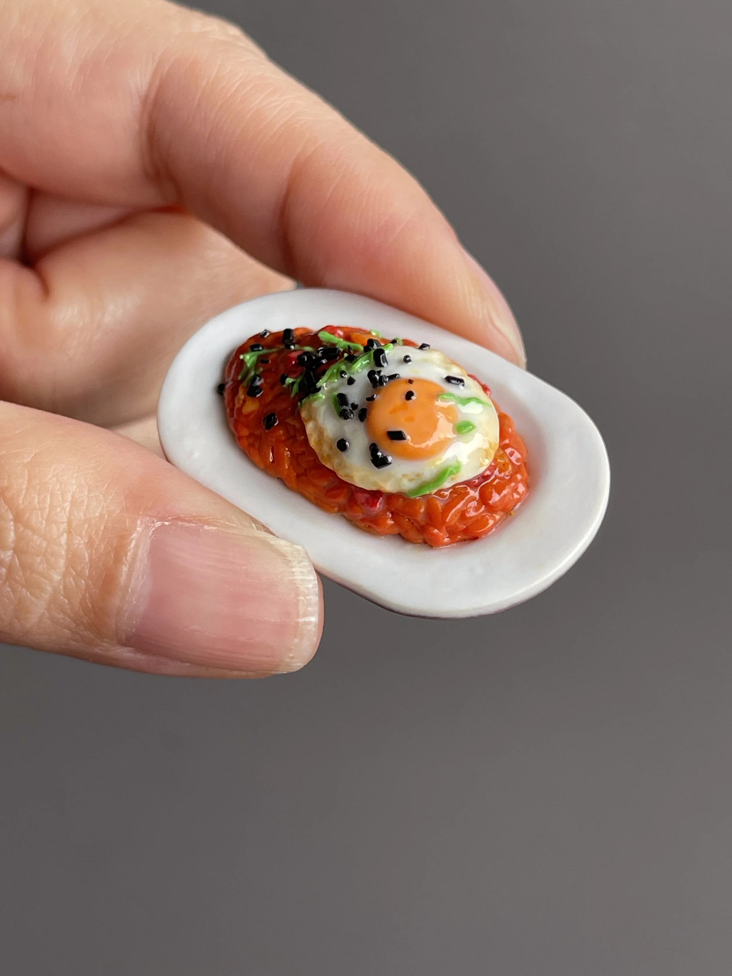 Fingers holding a miniature polymer clay kimchi fried rice topped with a fried egg, green onion, and sesame seeds atop a white plate.