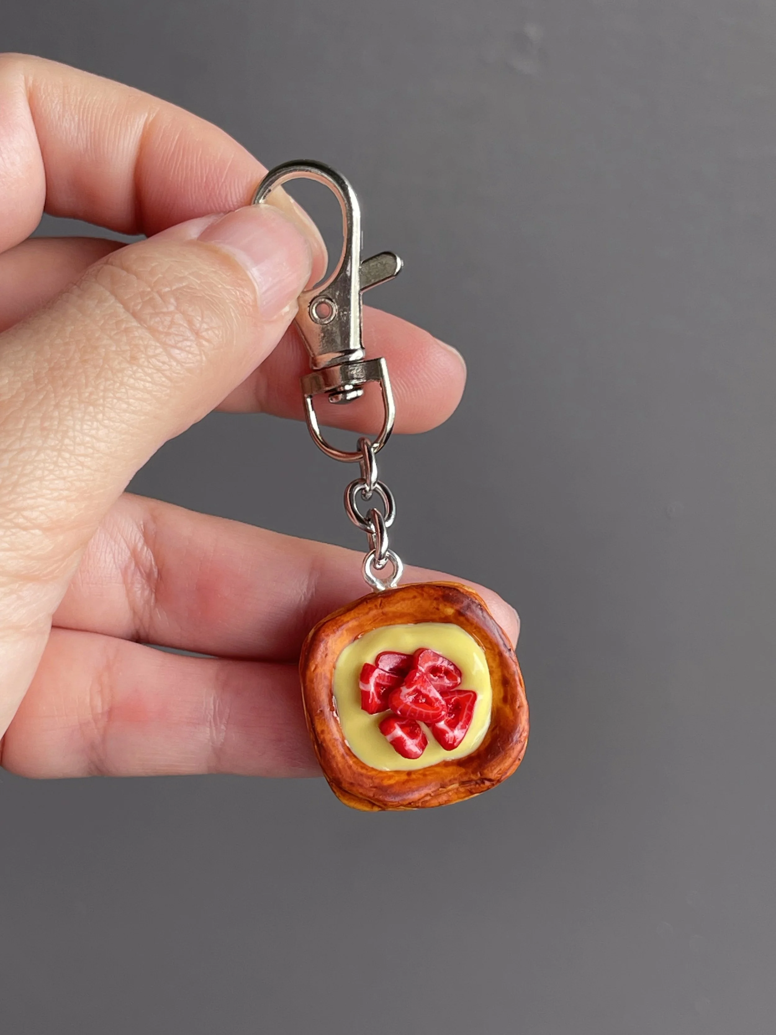 A hand holding a miniature danish pastry with yellow custard and strawberry slices keychain.
