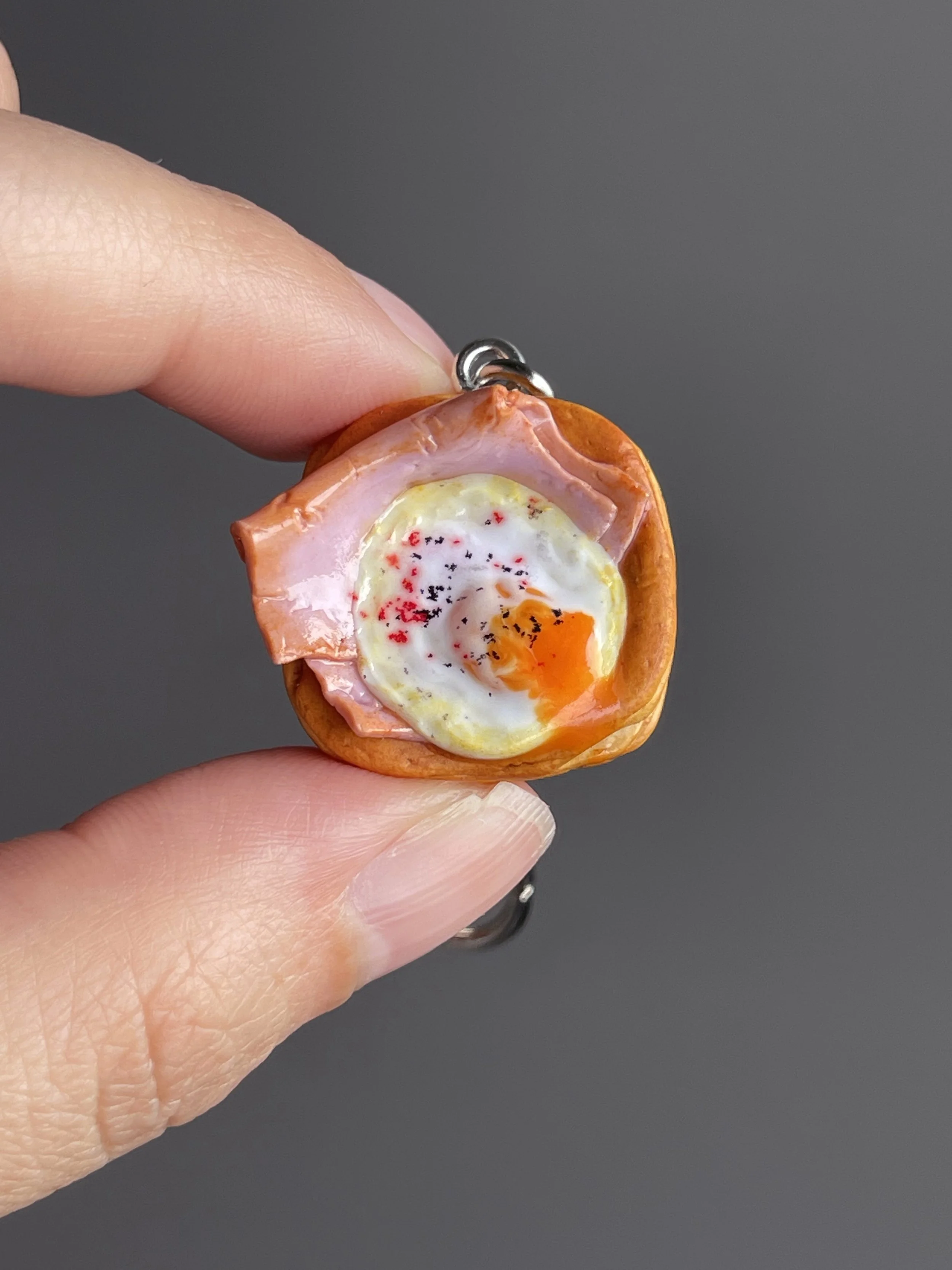 A hand holding a miniature polymer clay danish keychain with a piece of ham and egg with runny egg yolk.