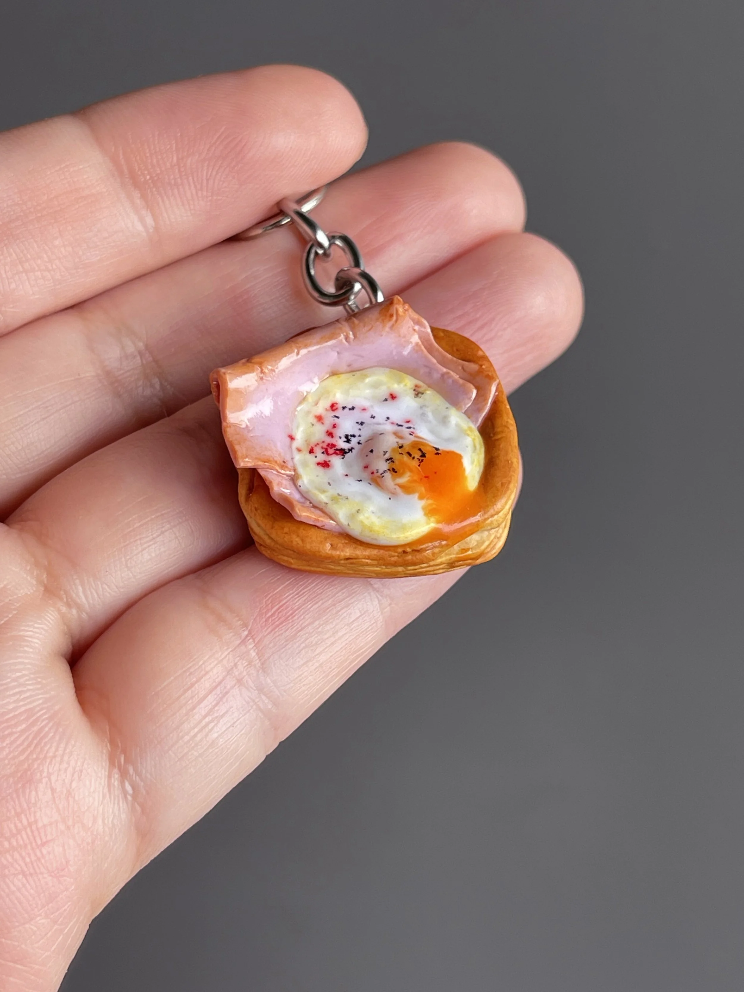 A hand holding a miniature polymer clay danish keychain with a piece of ham and egg with runny egg yolk.