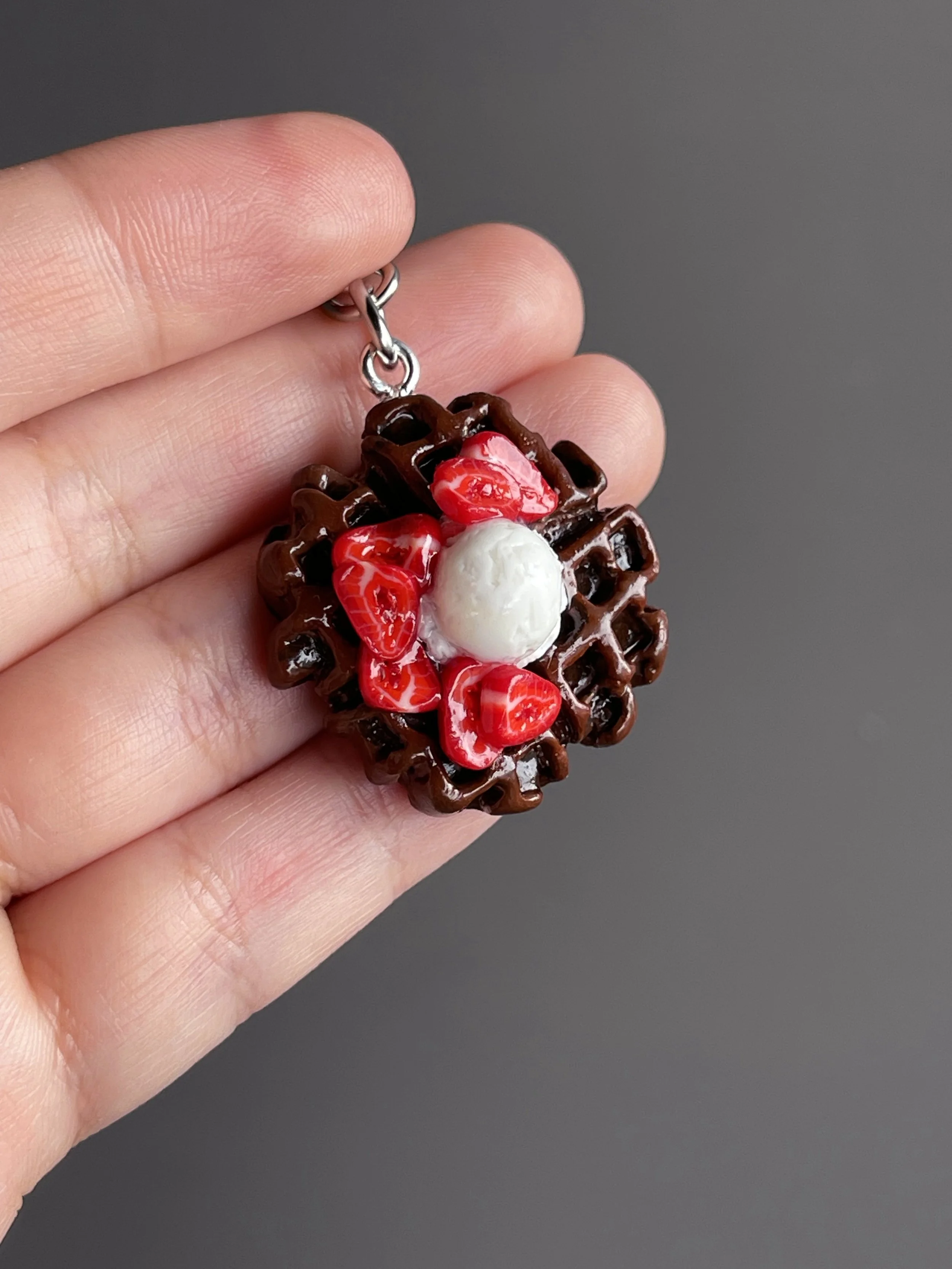 A hand holding a miniature polymer clay chocolate waffle keychain with strawberry slices & ice cream toppings.