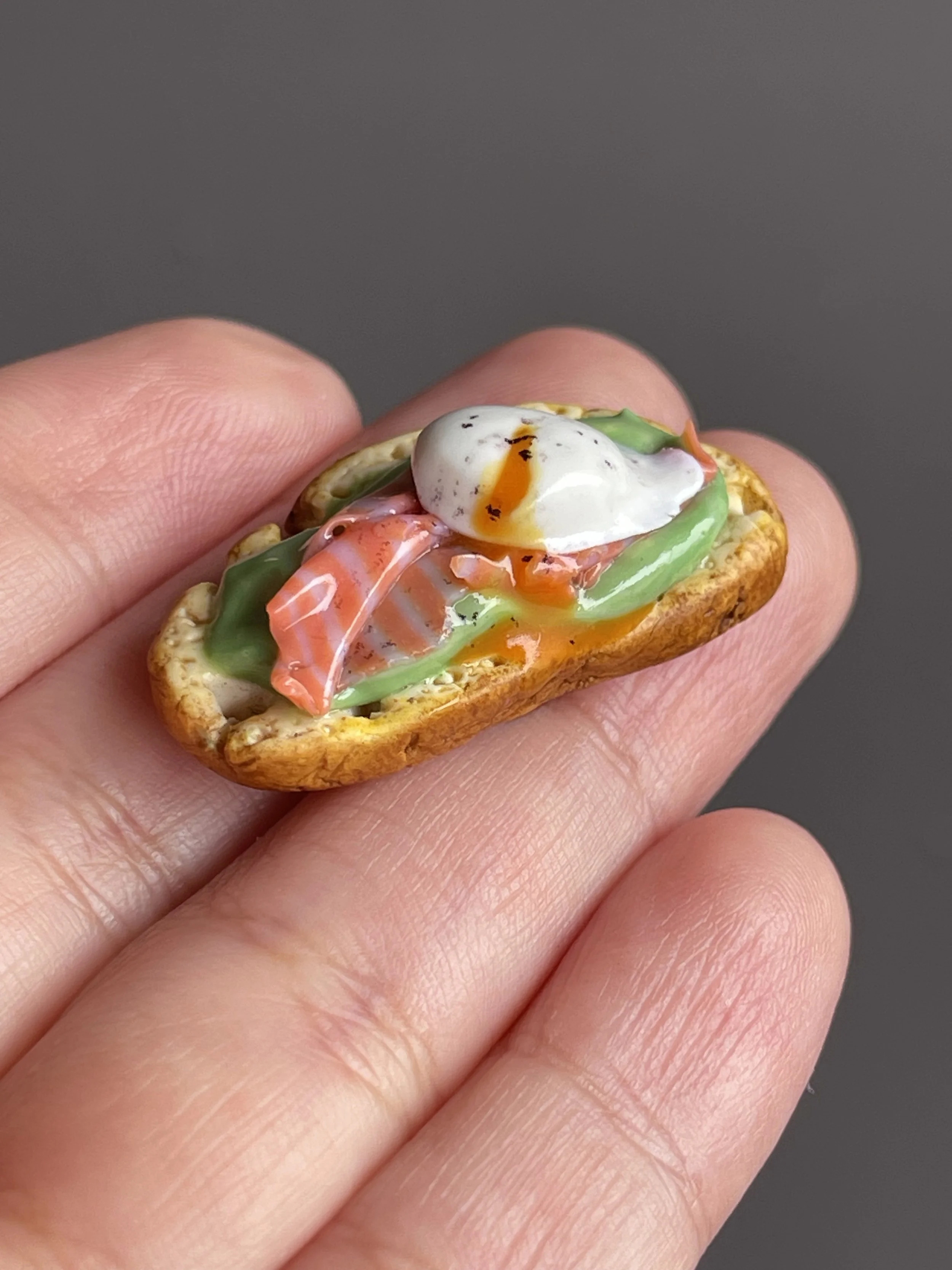 A hand holding a miniature polymer clay avocado toast magnet with smoked salmon, avocado mash, and a runny poached egg.