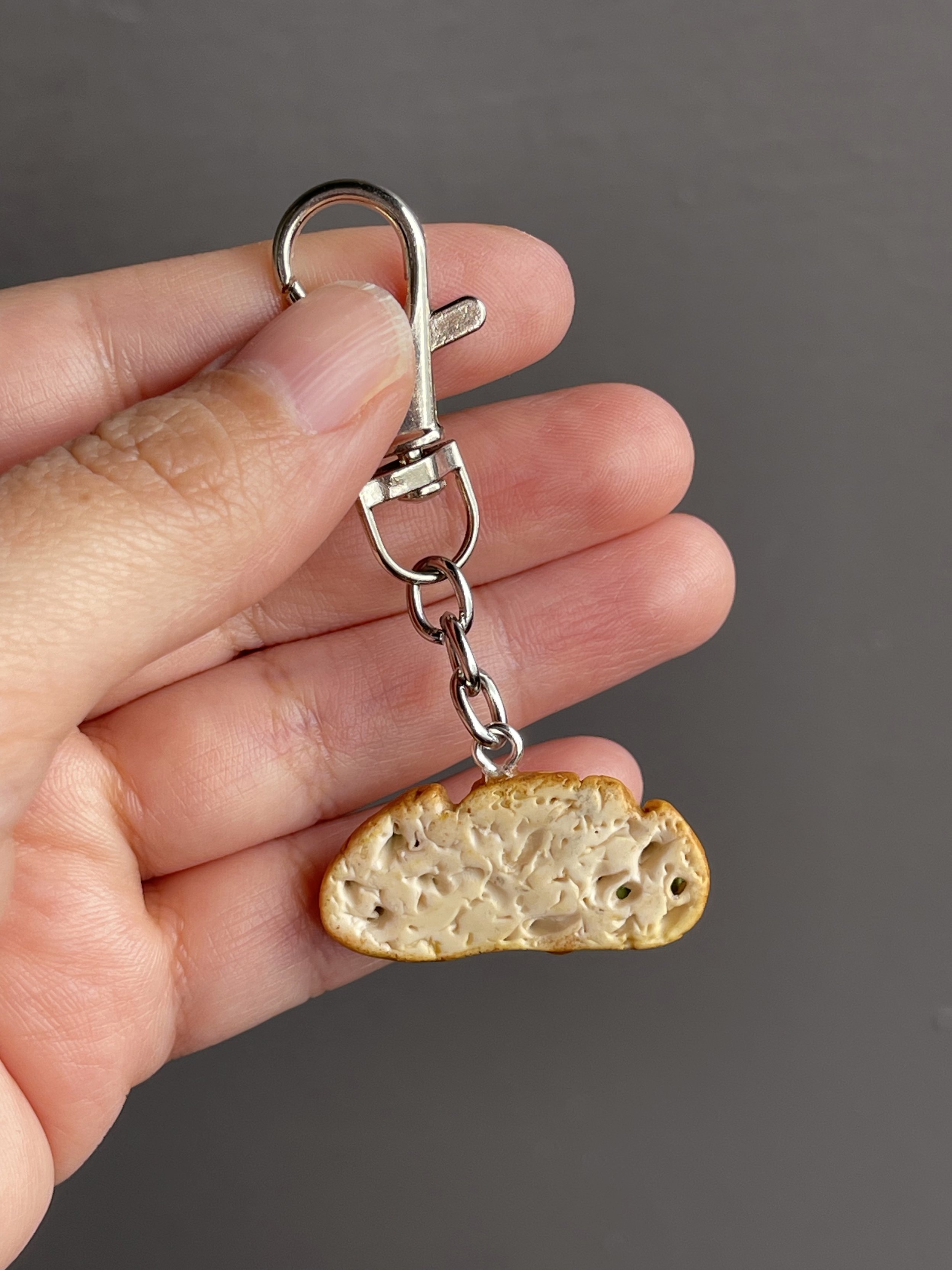 A hand holding a miniature polymer clay avocado toast keychain with smoked salmon, avocado mash, and a runny poached egg.