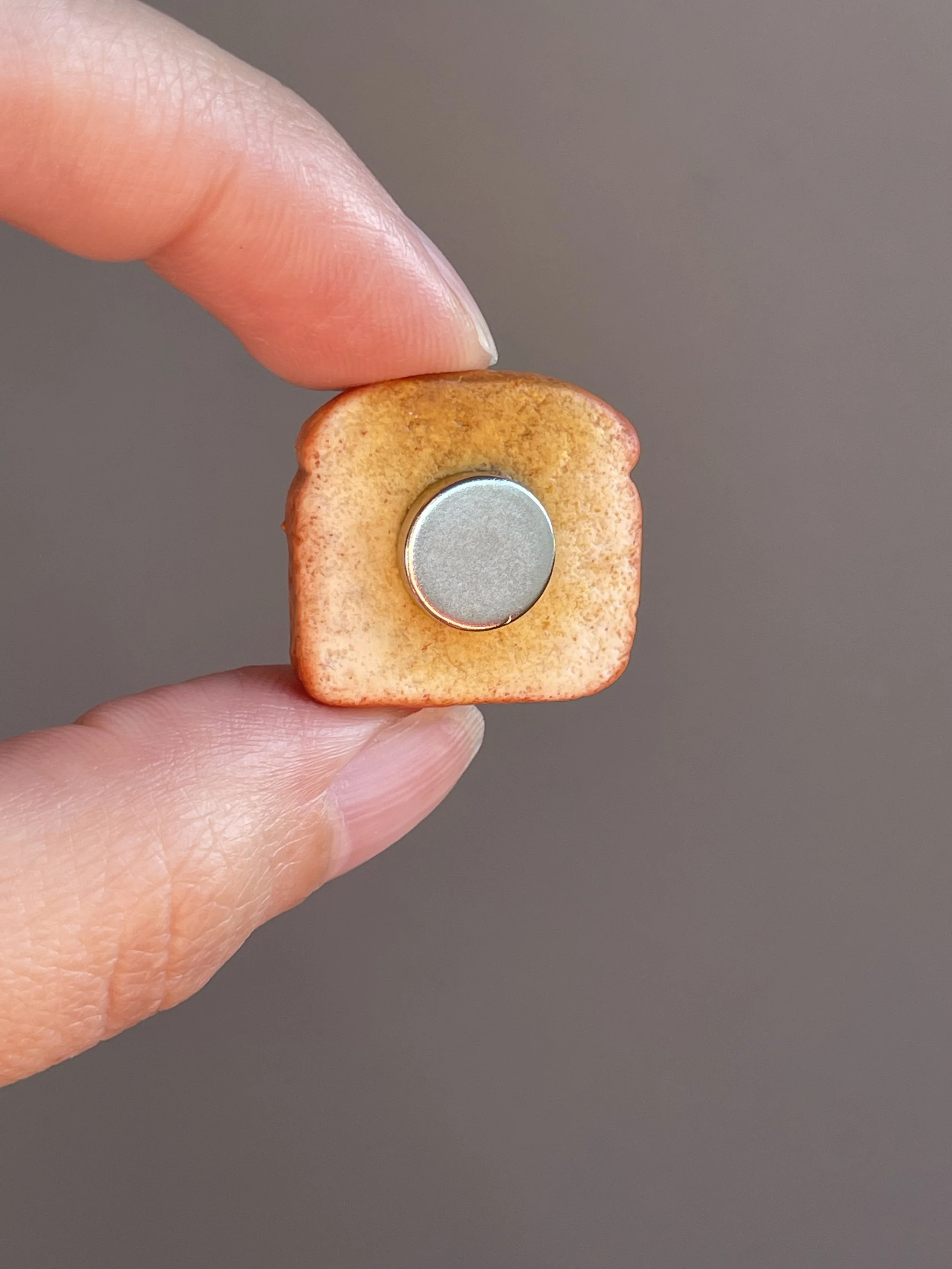 Fingers holding a miniature polymer clay hazelnut spread and sliced strawberries toast magnet showing the back.