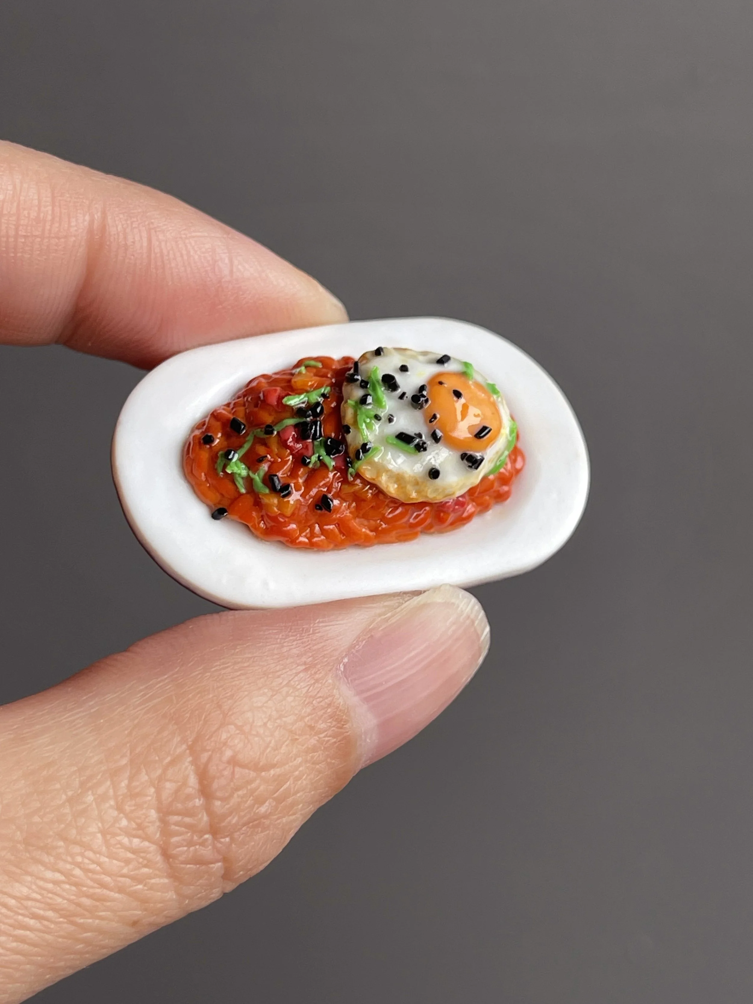 Fingers holding a miniature polymer clay kimchi fried rice topped with a fried egg, green onion, and sesame seeds atop a white plate.
