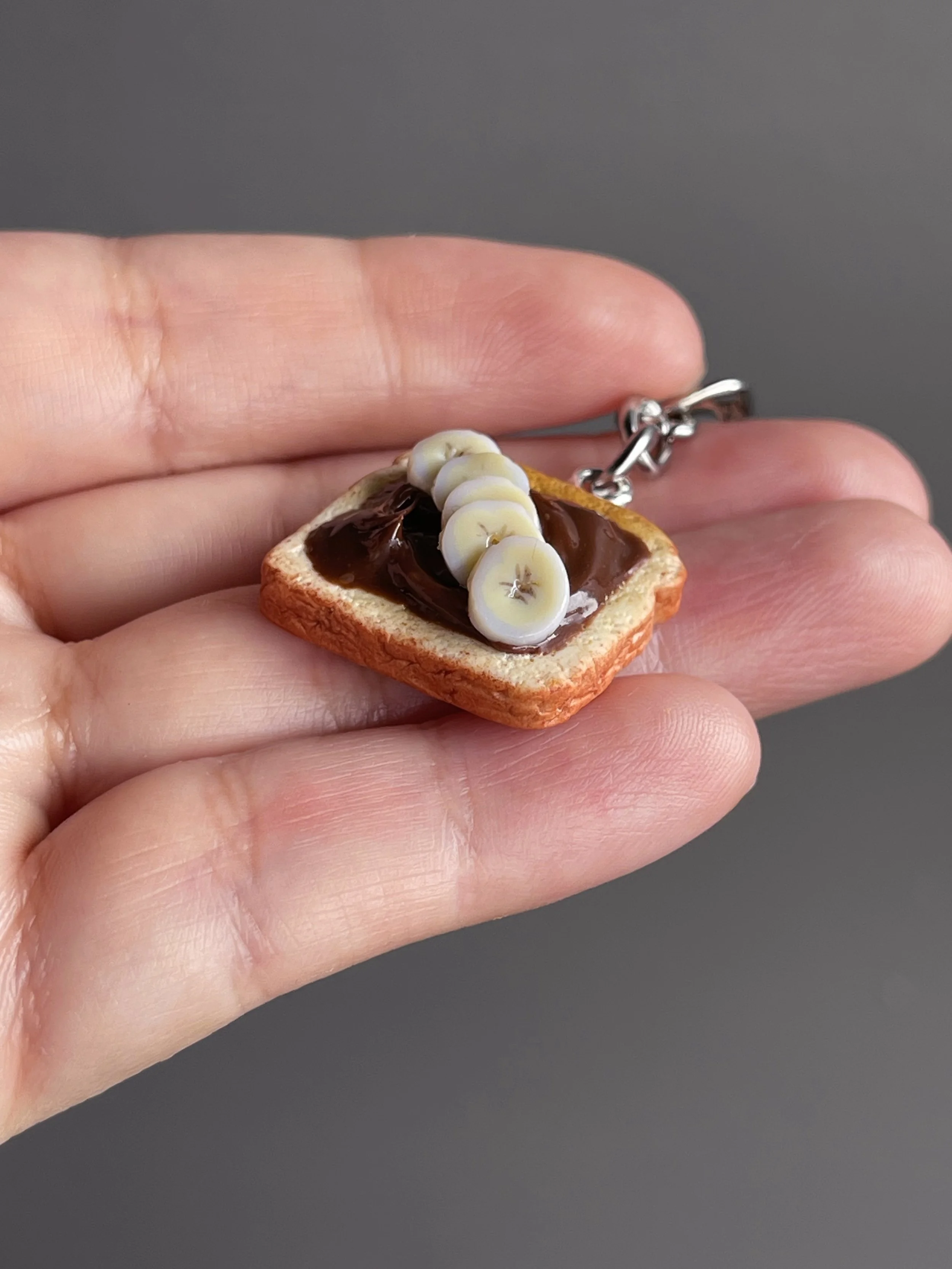 A hand holding a miniature polymer clay toast with hazelnut spread and banana slices.