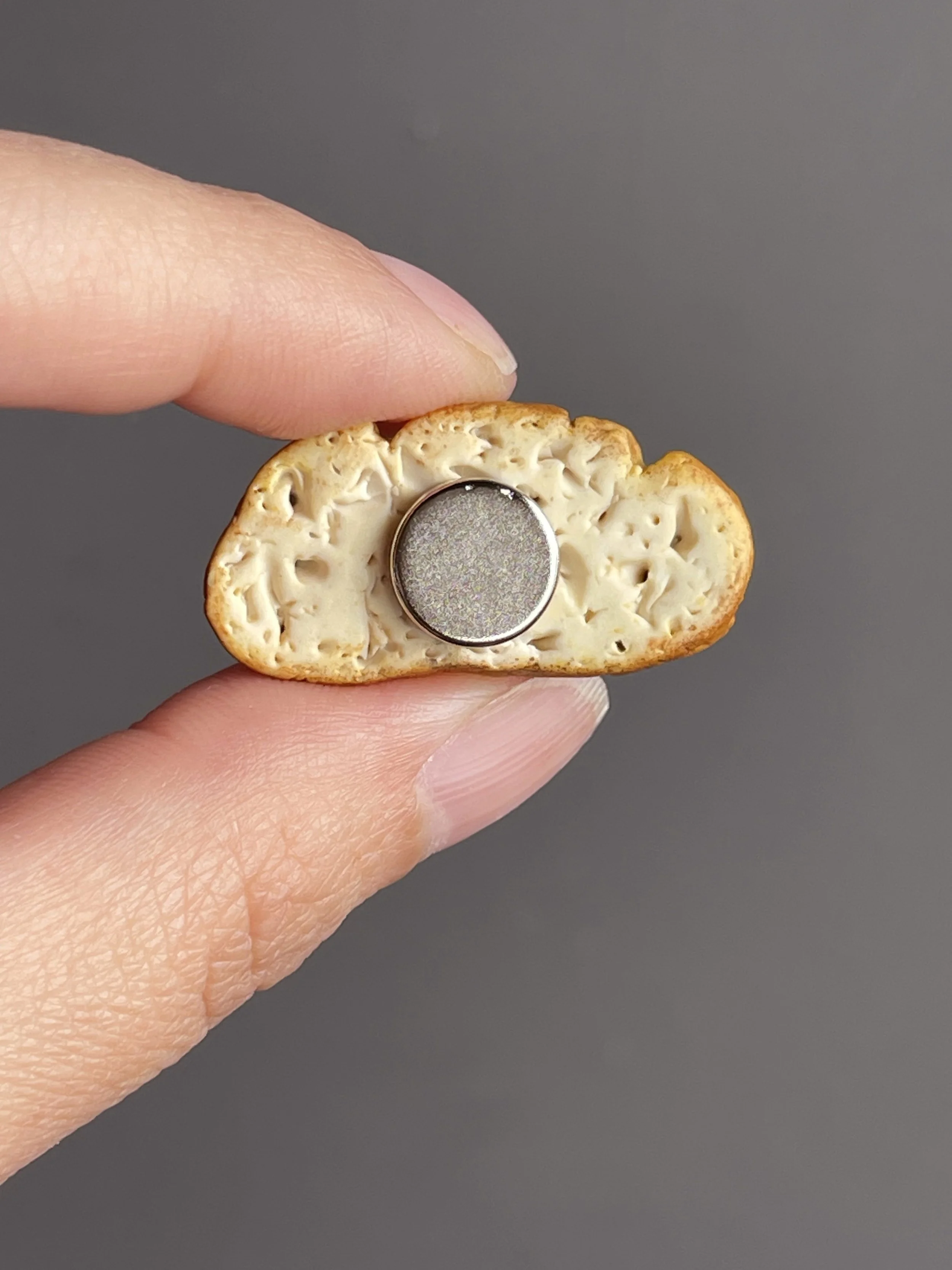 Fingers holding a miniature polymer clay avocado toast magnet with smoked salmon, avocado mash, and a runny poached egg showing the back.