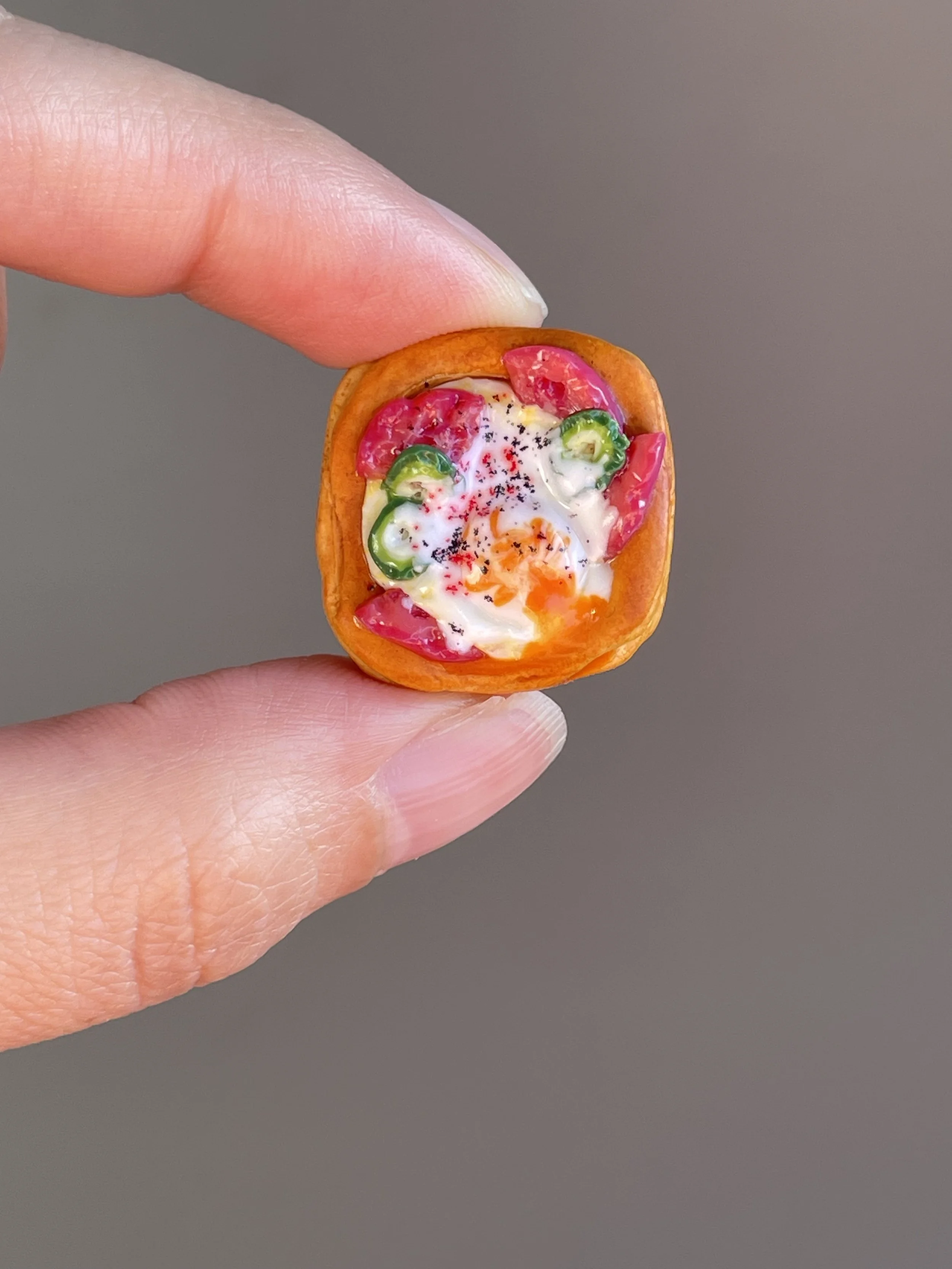 Fingers holding a miniature polymer clay egg, tomato, jalapeño breakfast danish magnet.