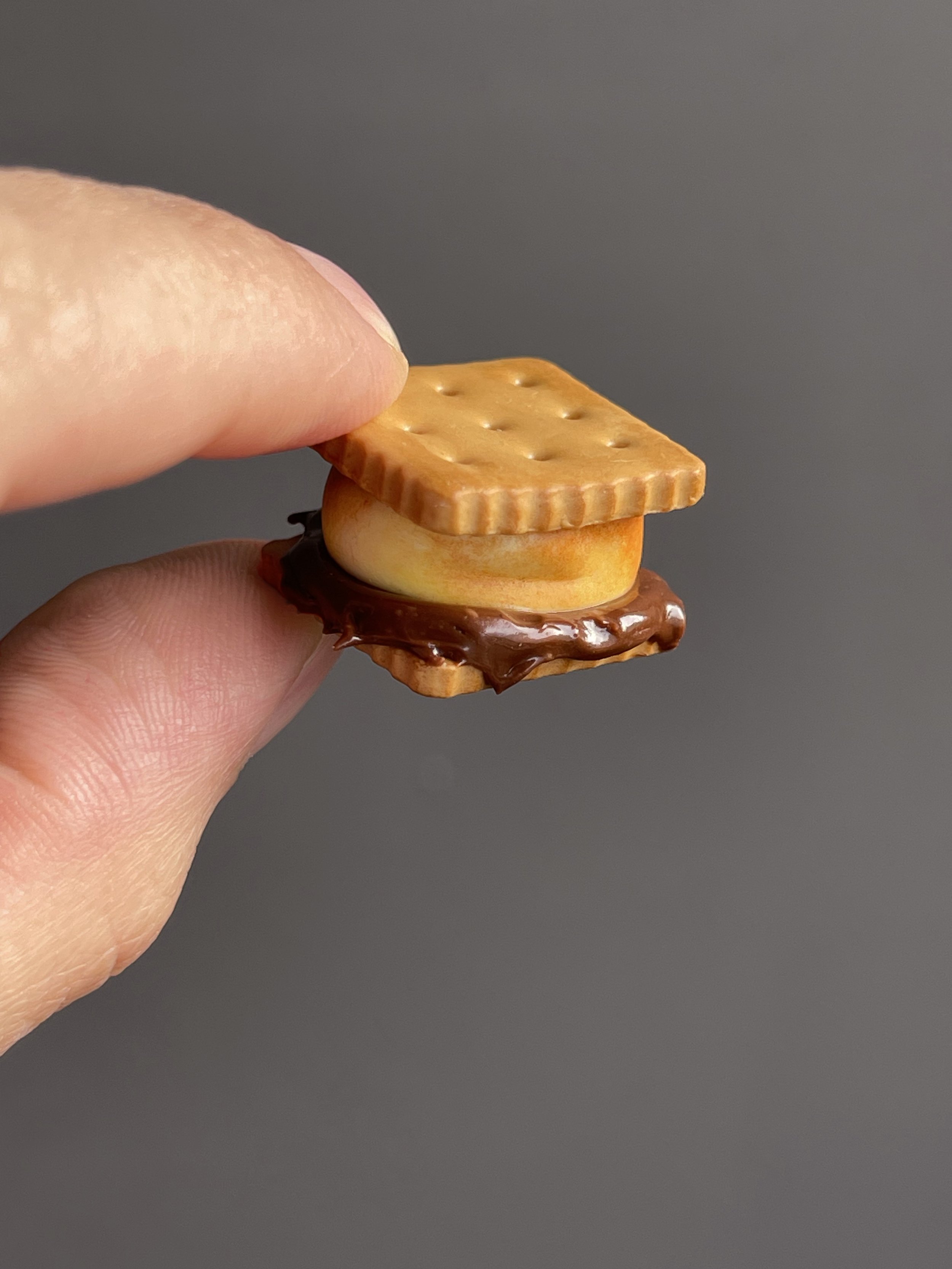 Fingers holding a miniature polymer clay s’more magnet.