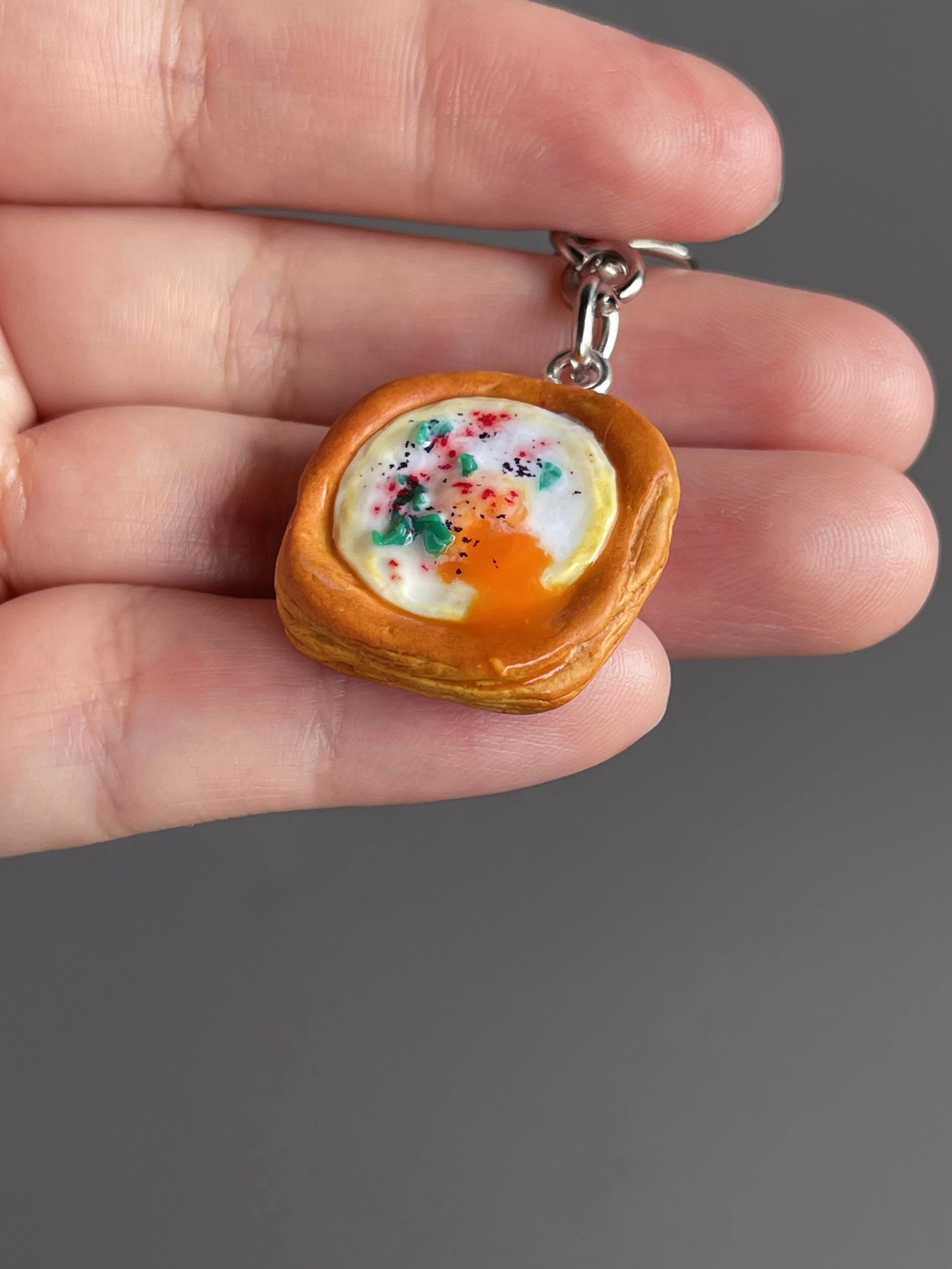 A hand holding a miniature polymer clay egg danish keychain with runny egg yolk.