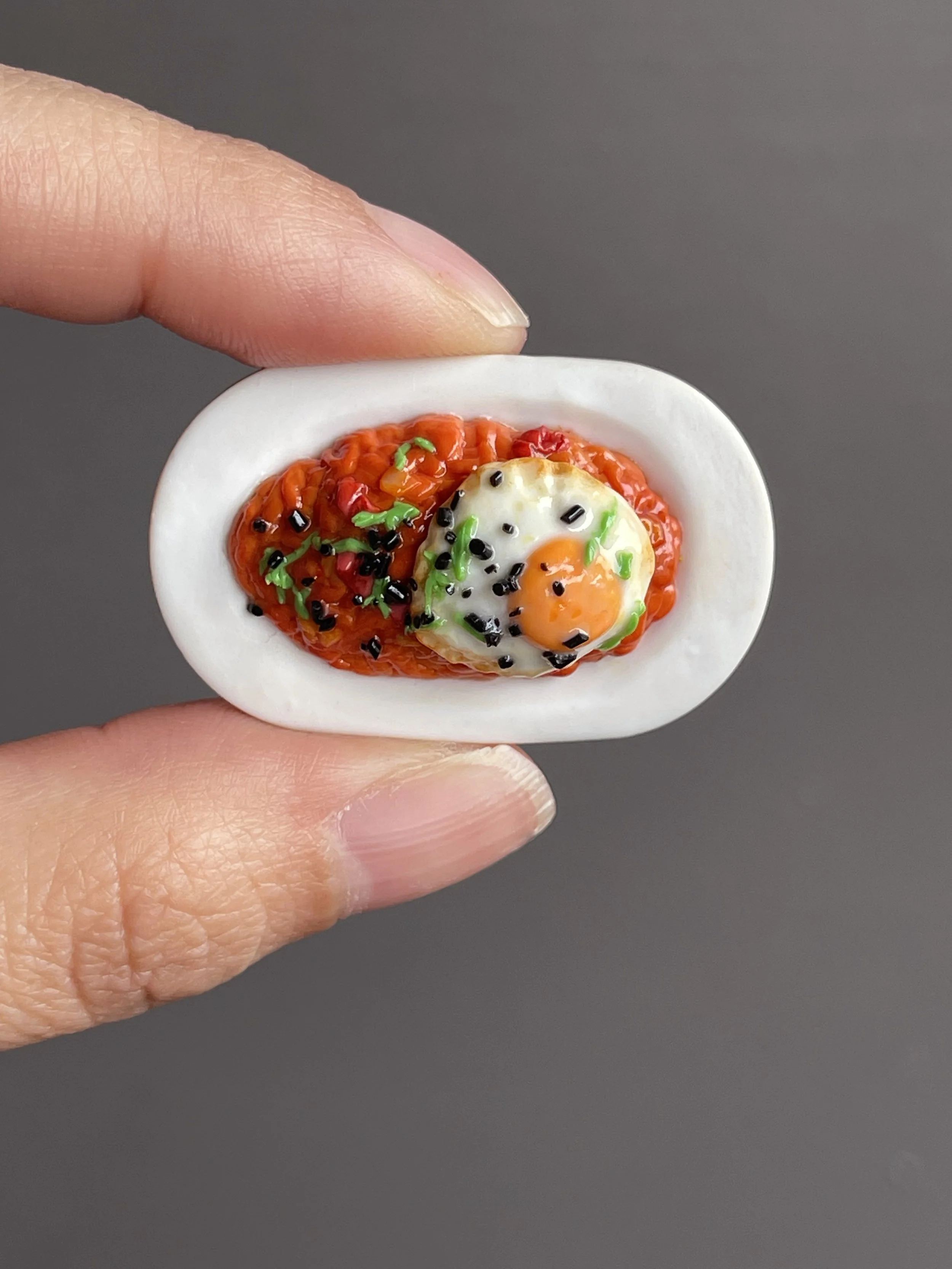 Fingers holding a miniature polymer clay kimchi fried rice topped with a fried egg, green onion, and sesame seeds atop a white plate.