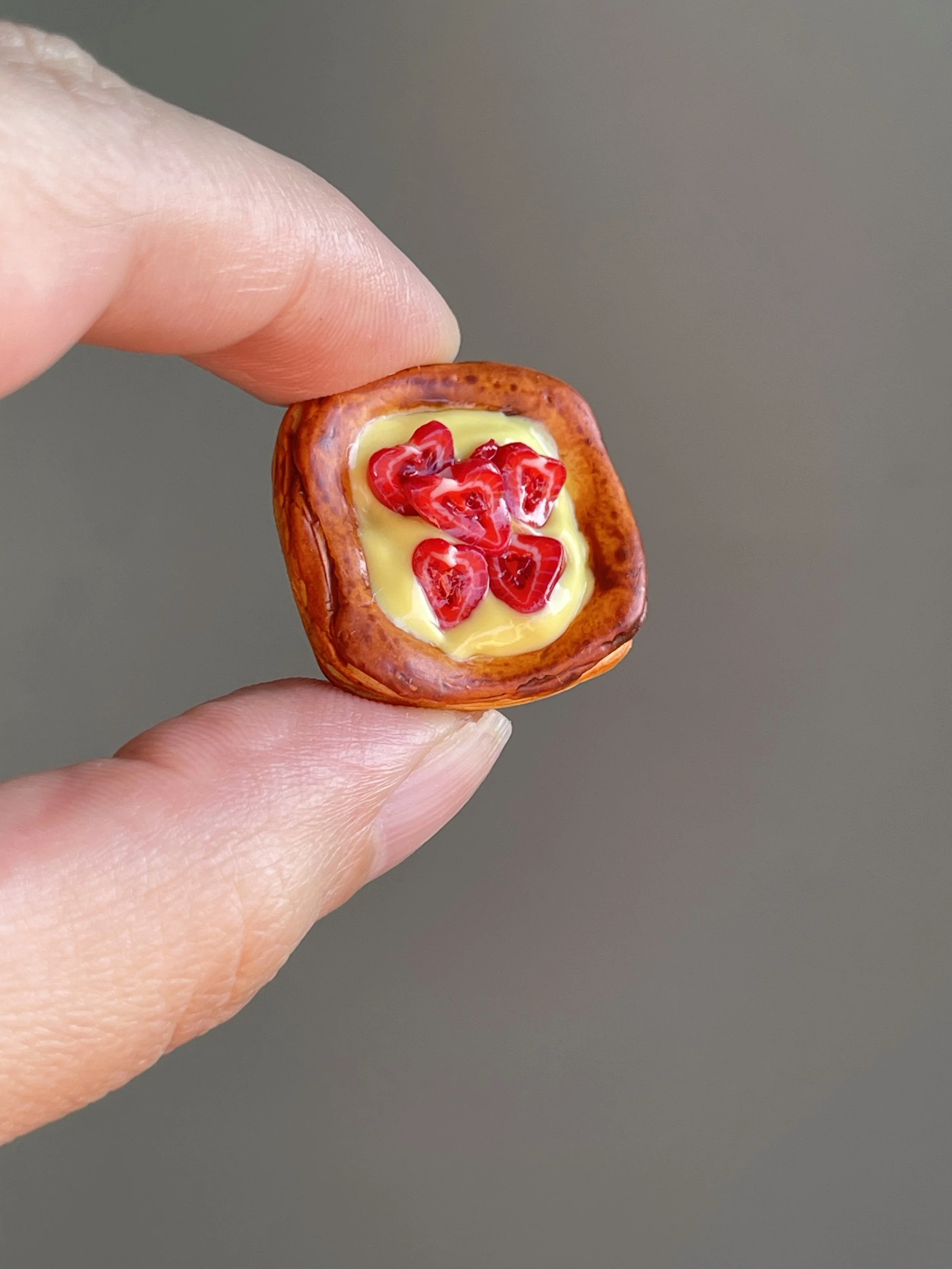 Fingers holding a miniature polymer clay danish with yellow custard and strawberry slices magnet.