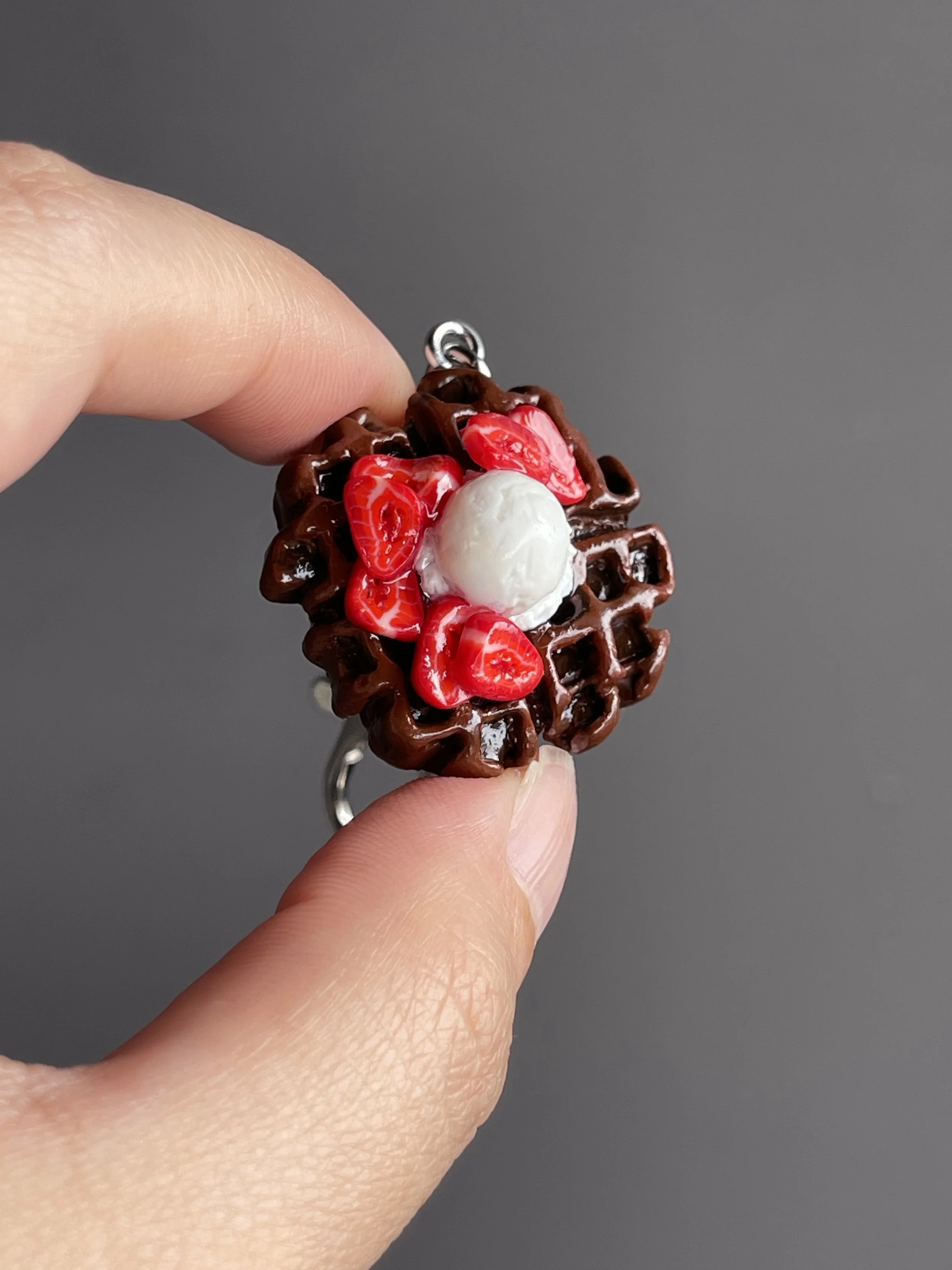 A hand holding a miniature polymer clay chocolate waffle keychain with strawberry slices & ice cream toppings.