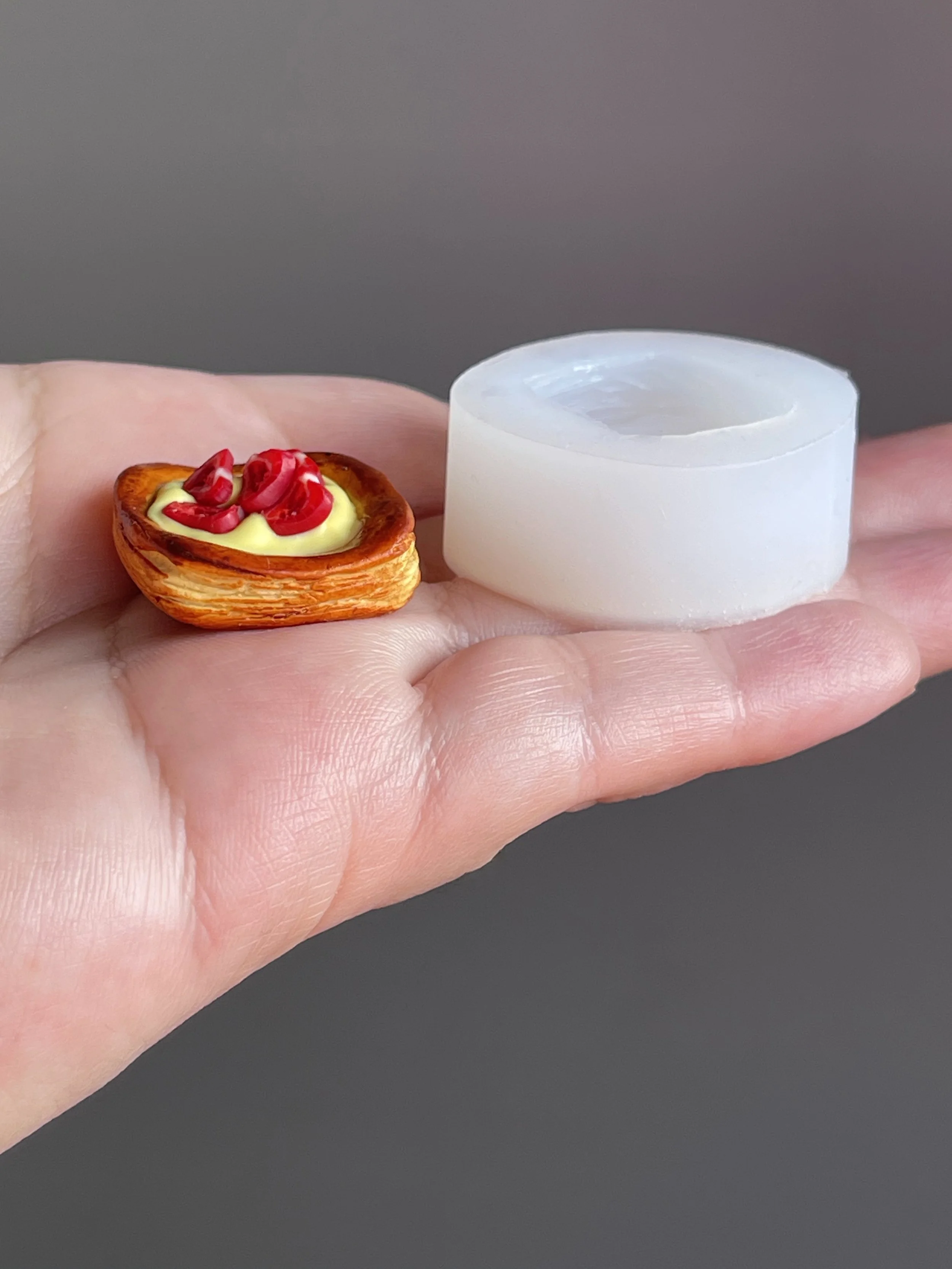 Hand holding a polymer clay danish with yellow cream and strawberry slices next to the clear silicone mold used to make it.