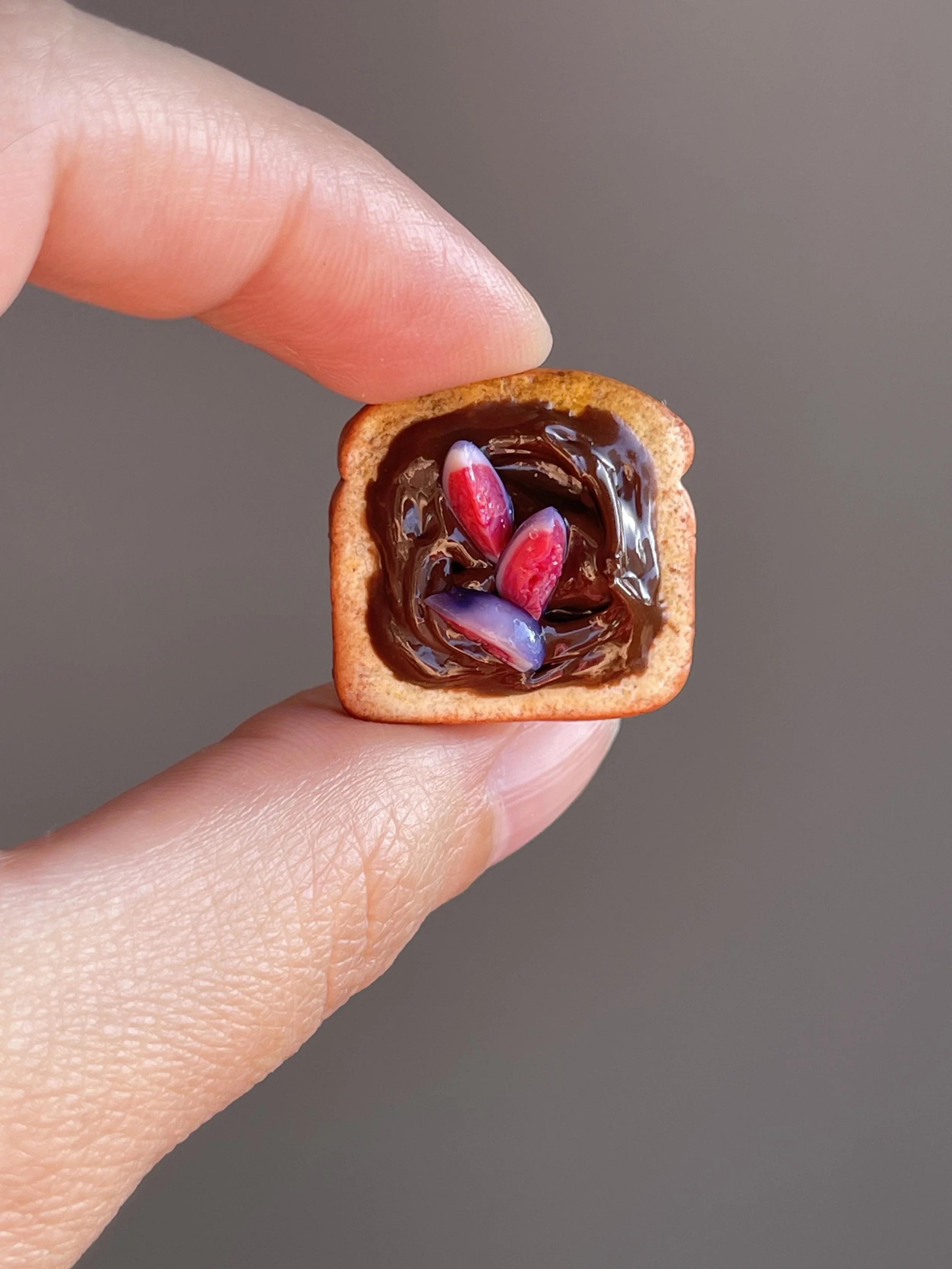 Fingers holding a miniature polymer clay hazelnut spread and figs toast magnet.
