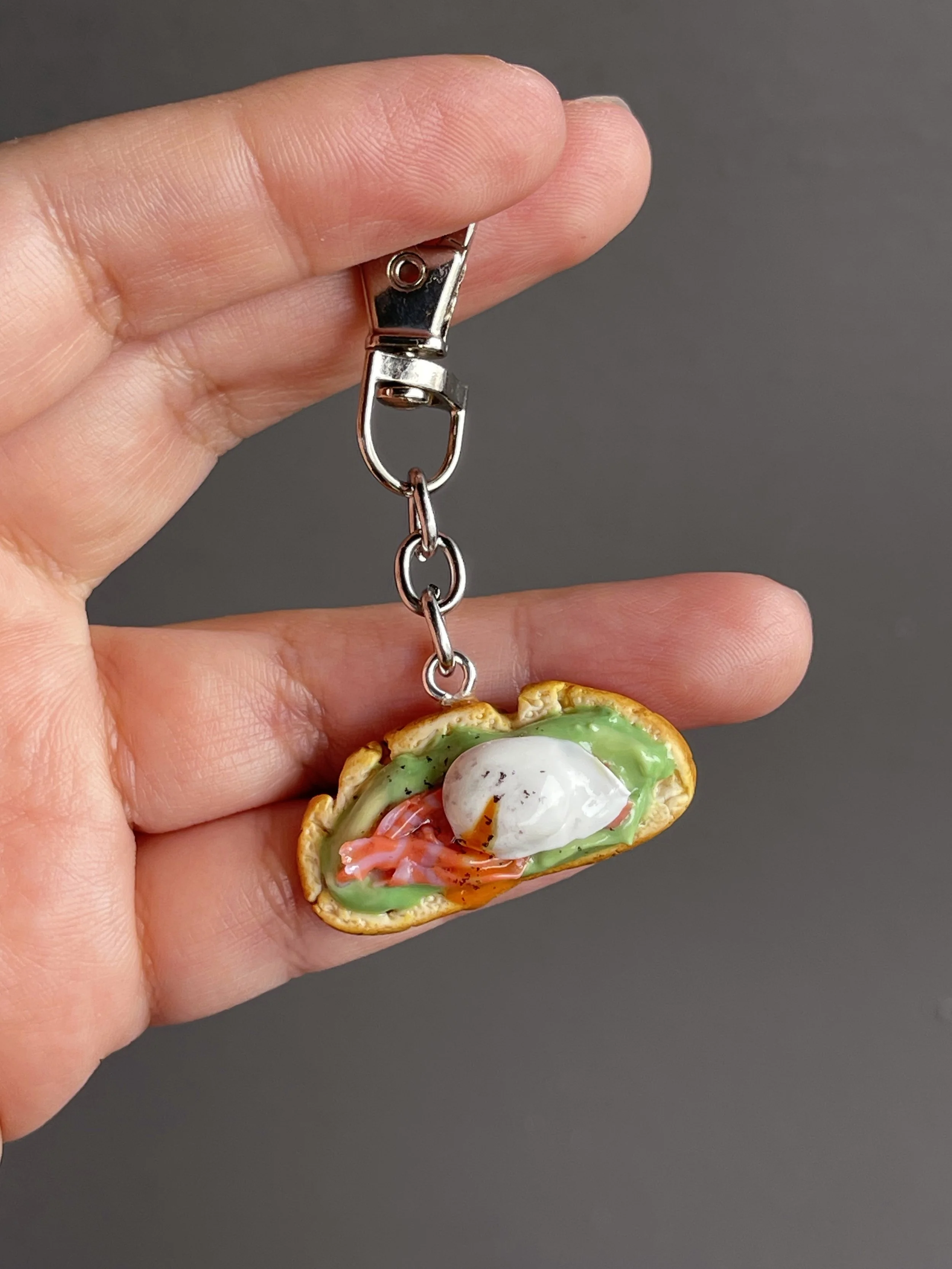A hand holding a miniature polymer clay avocado toast keychain with smoked salmon, avocado mash, and a runny poached egg.