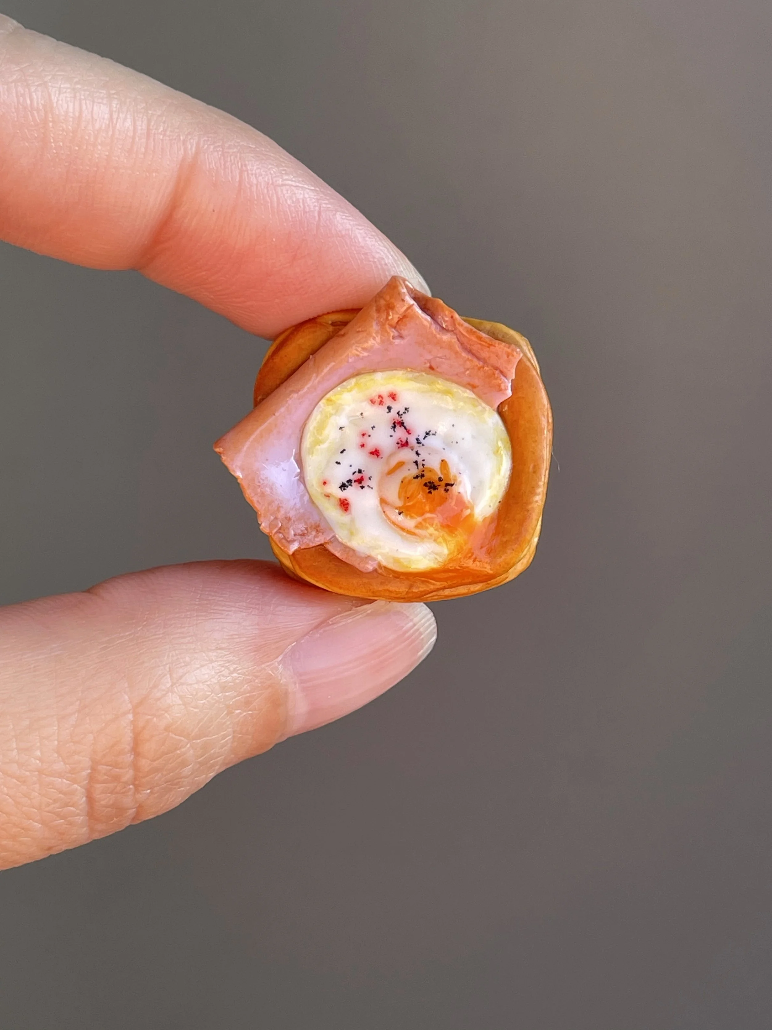 Fingers holding a miniature realistic polymer clay ham and egg danish magnet.