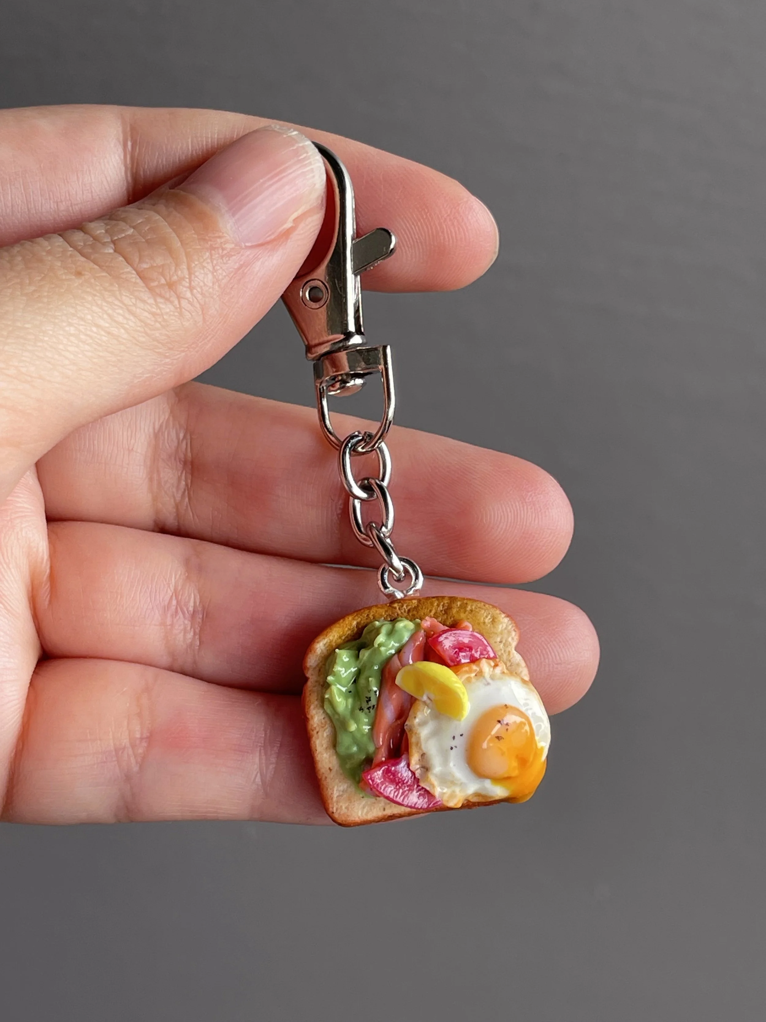 A hand holding a miniature polymer clay avocado, smoked salmon, tomato toast keychain with runny egg yolk and lemon slice garnish.