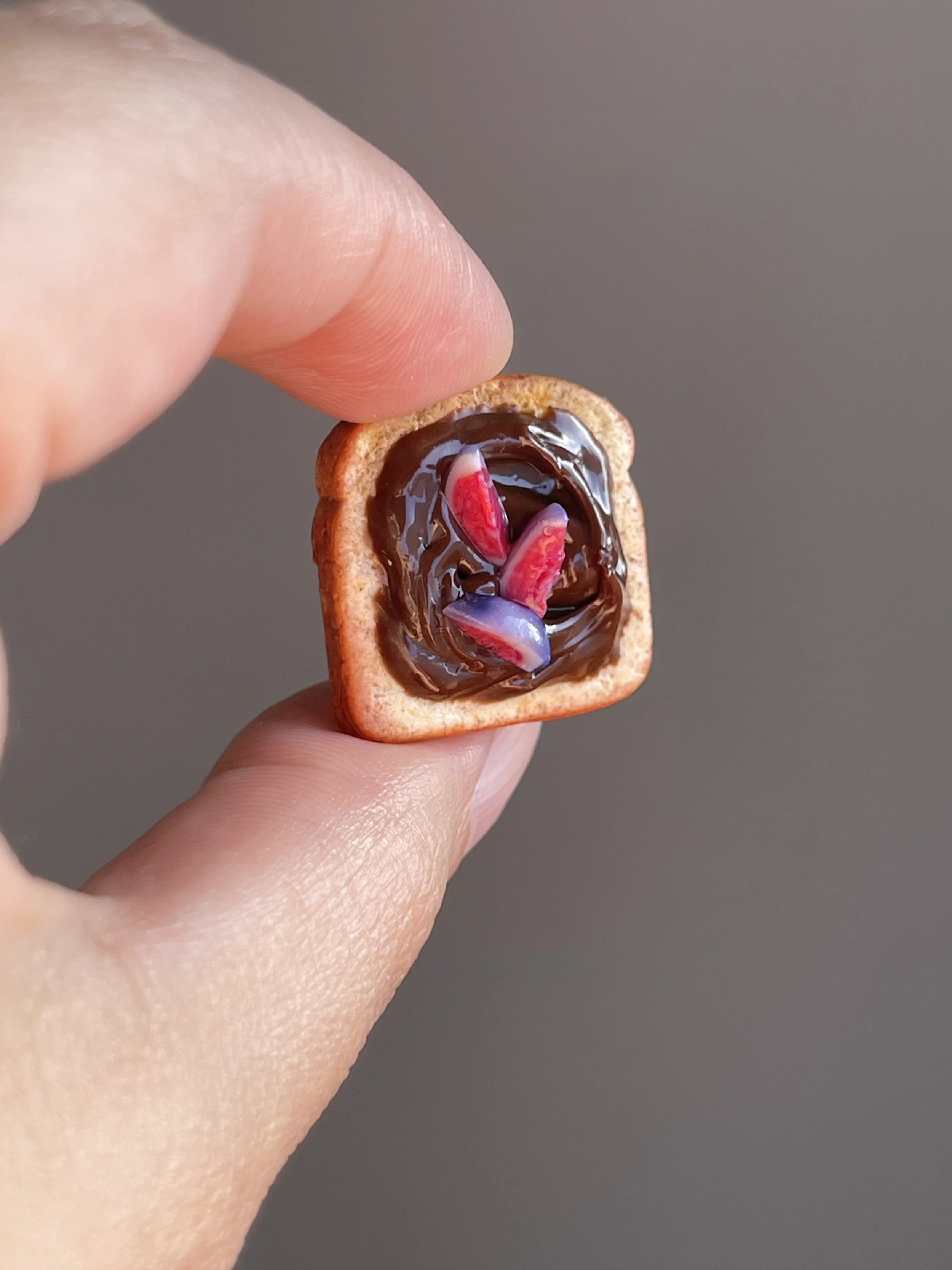 Fingers holding a miniature polymer clay hazelnut spread and figs toast magnet.