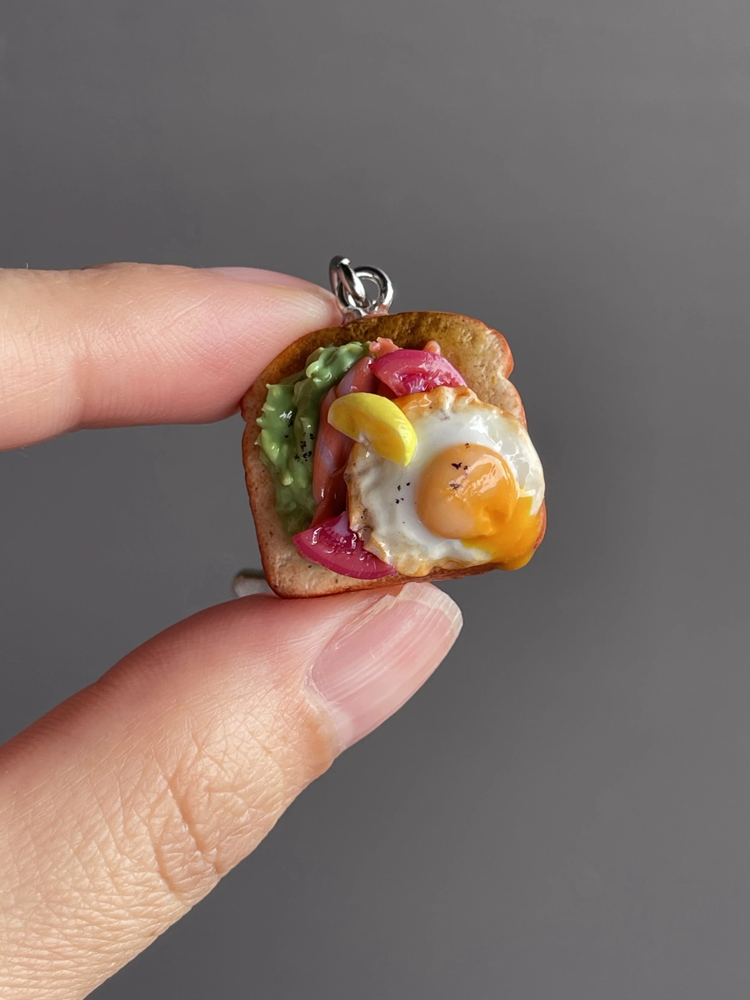A hand holding a miniature polymer clay avocado, smoked salmon, tomato toast keychain with runny egg yolk and lemon slice garnish.