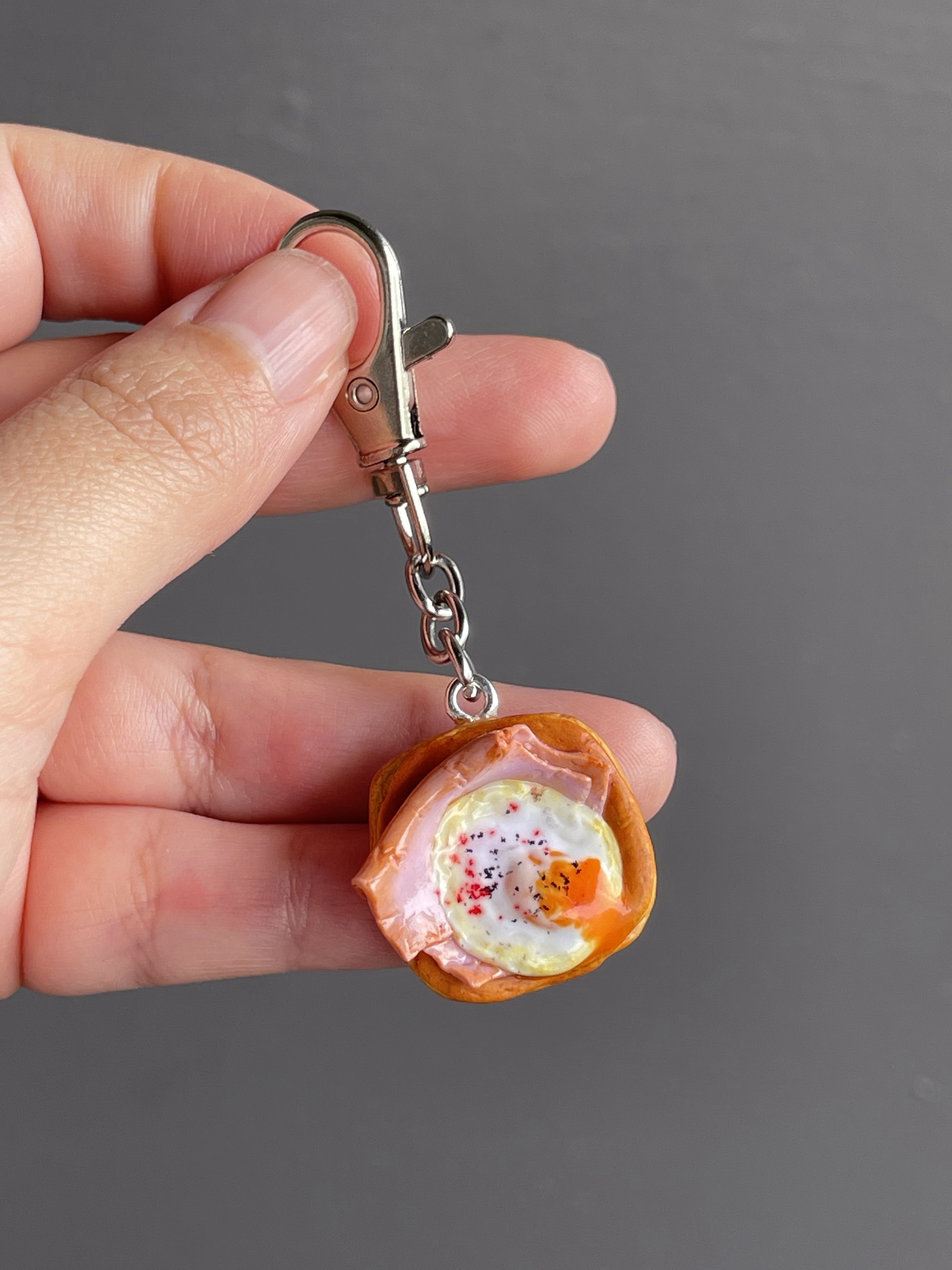 A hand holding a miniature polymer clay danish keychain with a piece of ham and egg with runny egg yolk.
