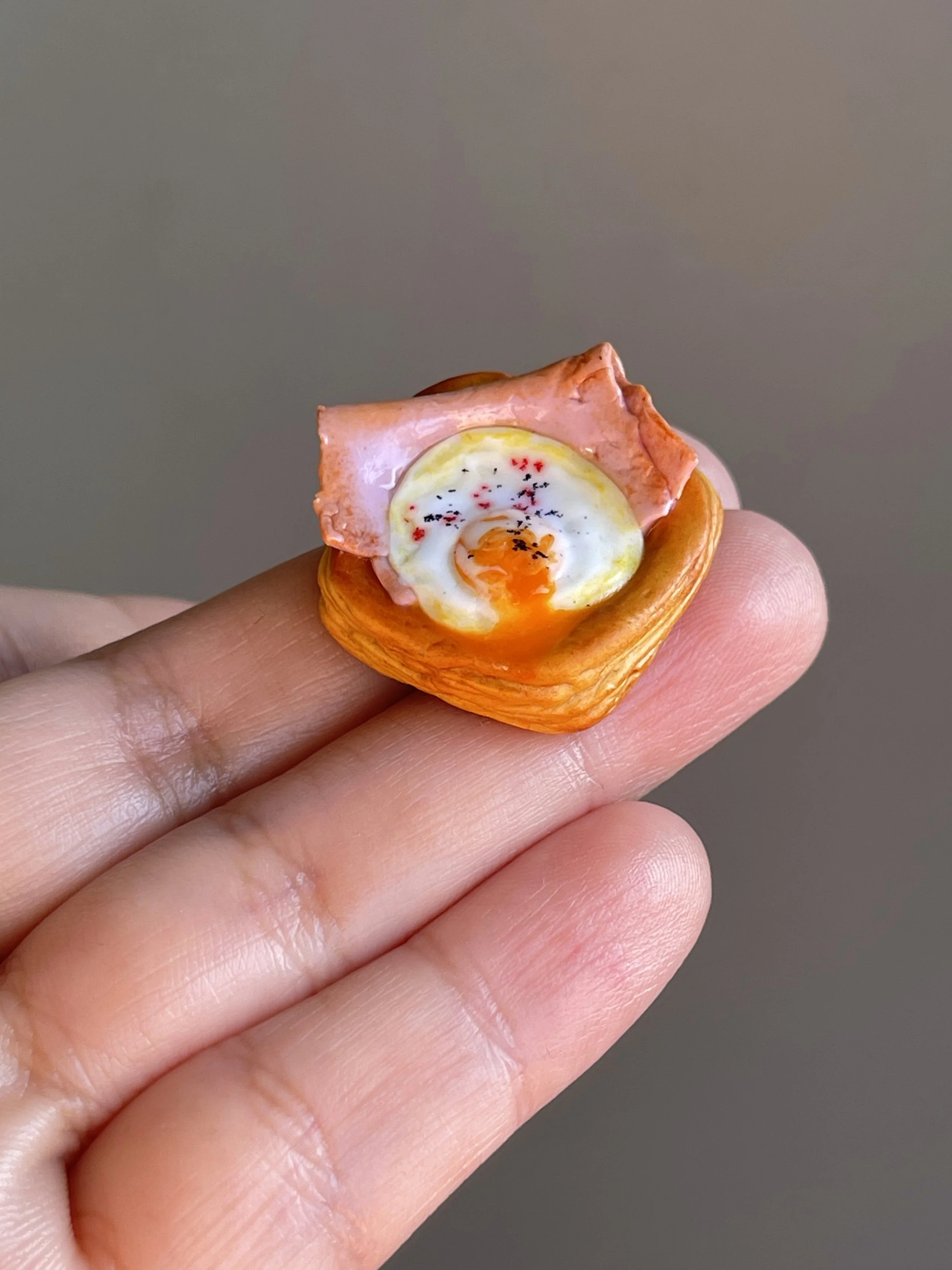 A hand holding a miniature realistic polymer clay ham and egg danish magnet.