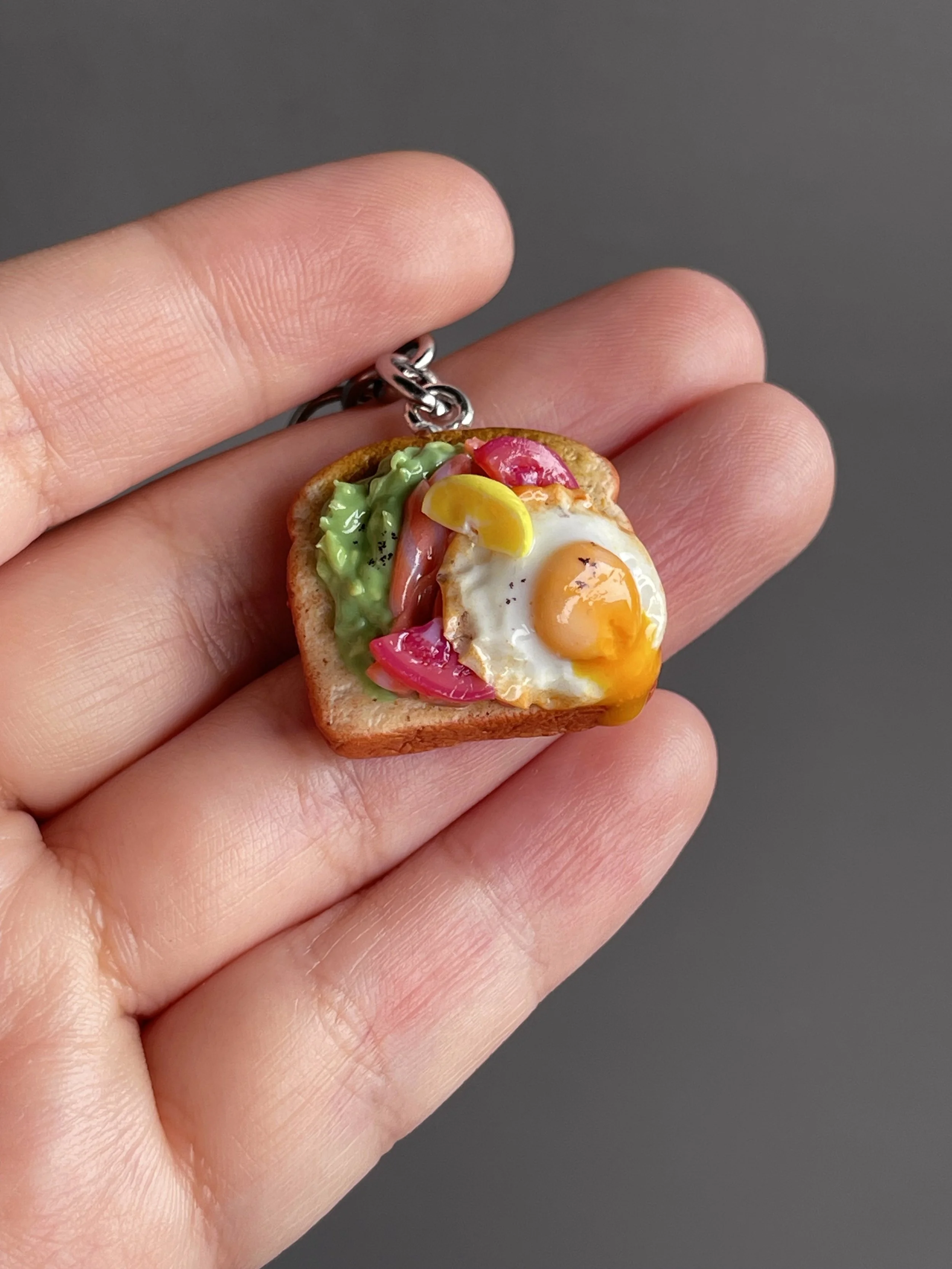 A hand holding a miniature polymer clay avocado, smoked salmon, tomato toast keychain with runny egg yolk and lemon slice garnish.